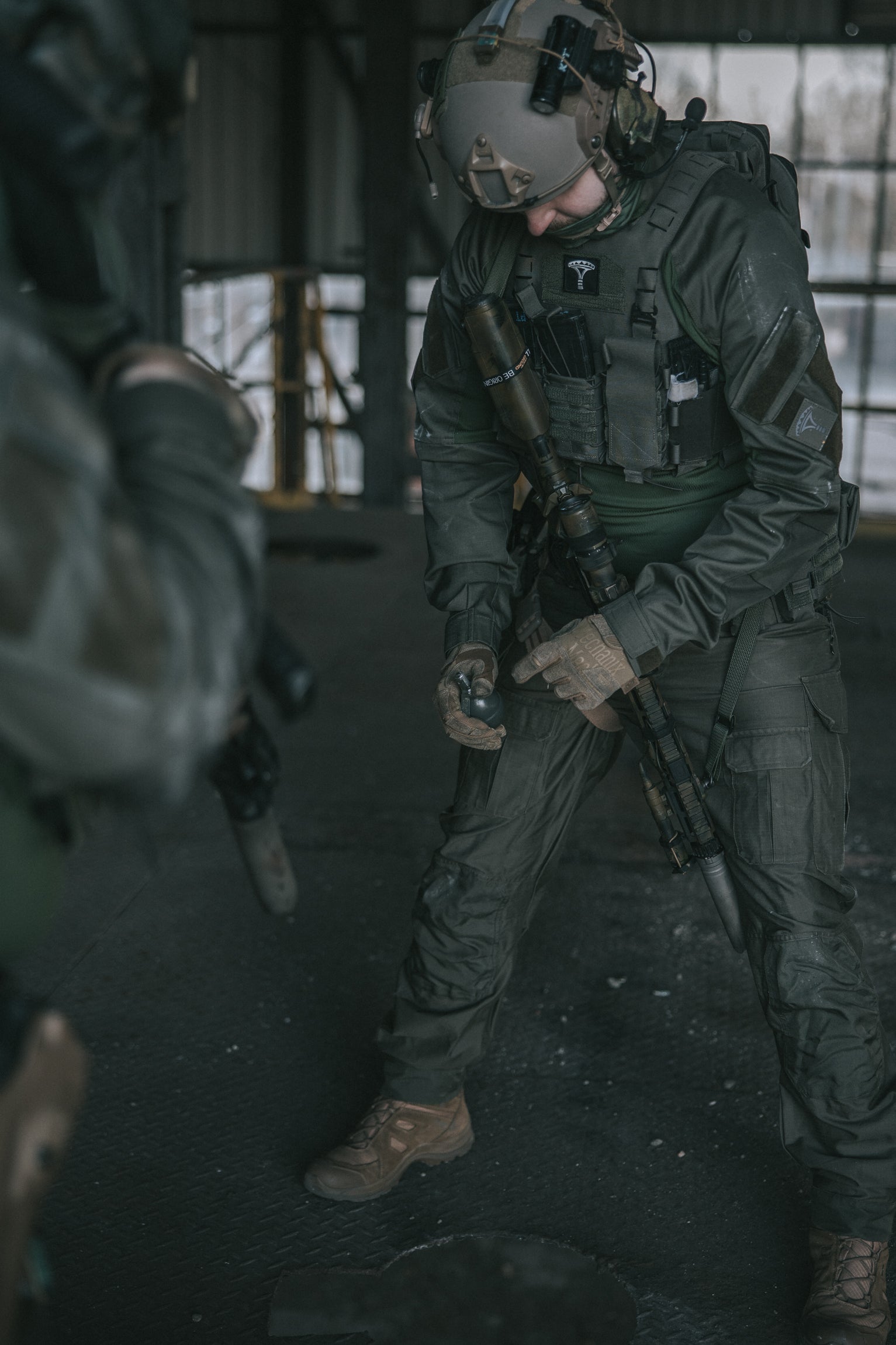 Person in tactical gear holding hand grenade and a rifle in an industrial setting