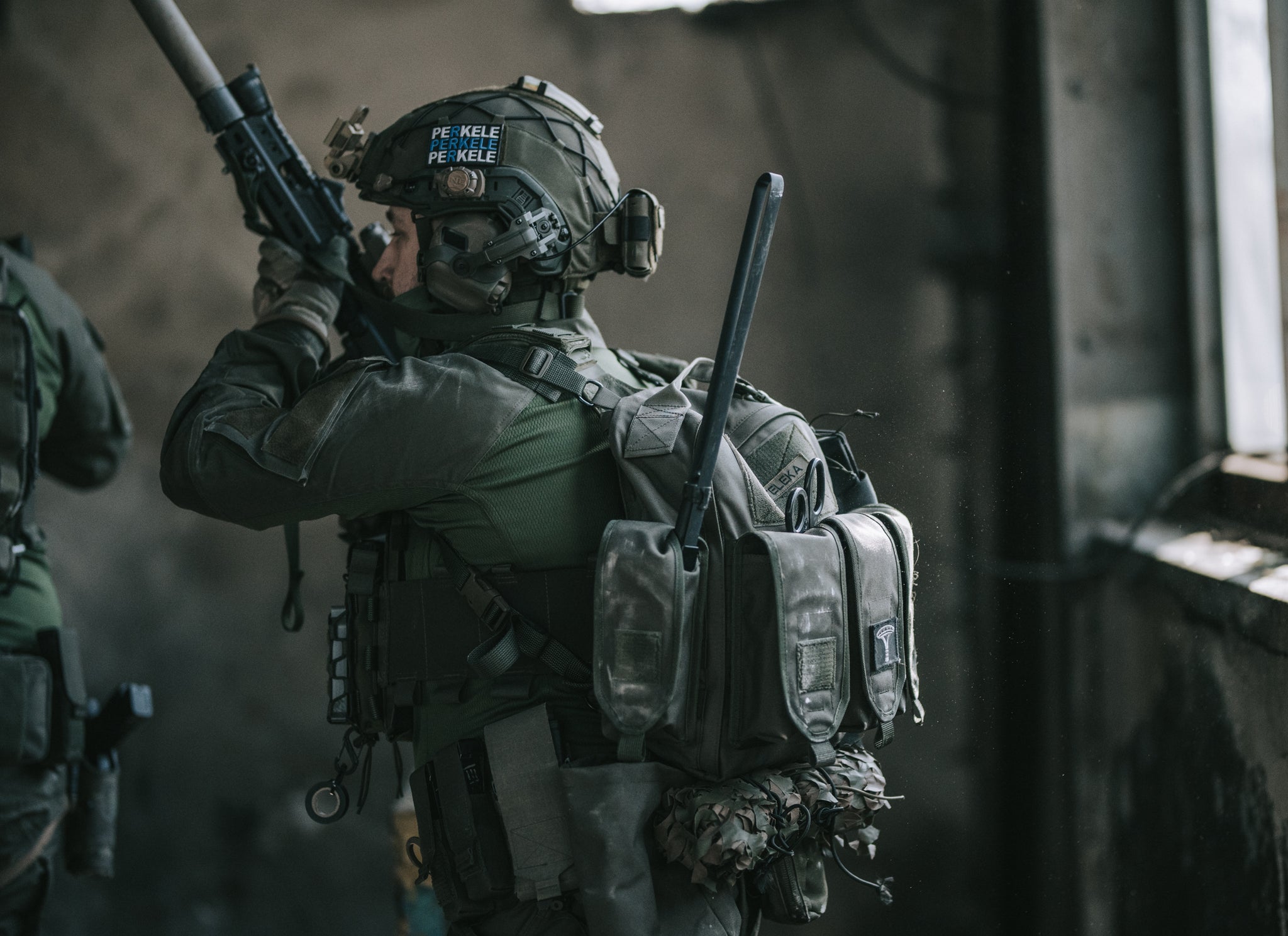 Soldier in tactical gear wearing a green combat shirt, green backpack and combat helmet holding a rifle in a dimly lit room