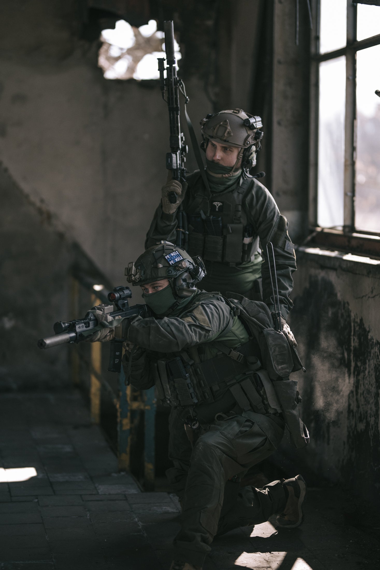 Two soldiers in tactical gear inside a dimly lit building.