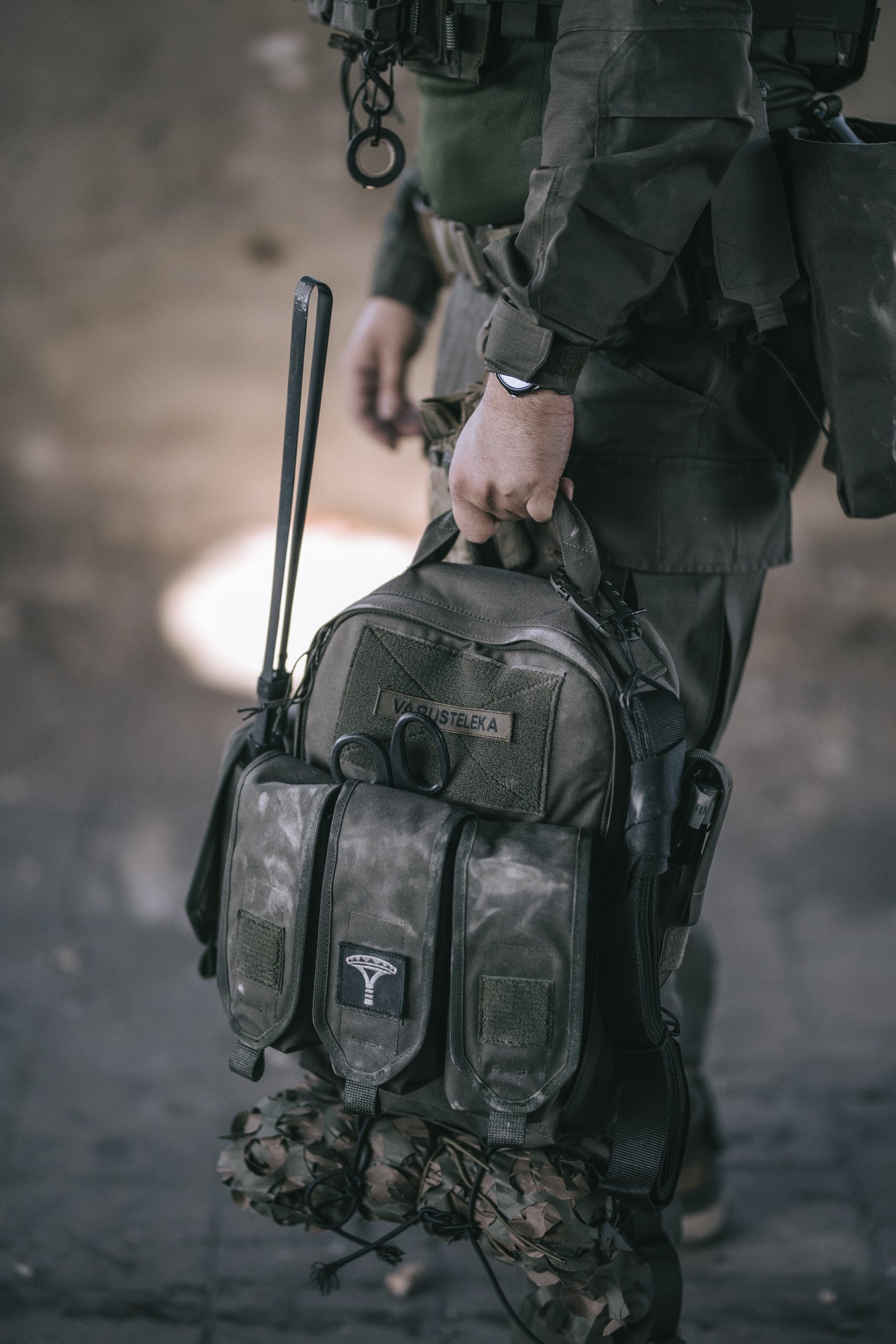 Person holding a tactical backpack with a blurred background. The person is dressed in green combat clothing.