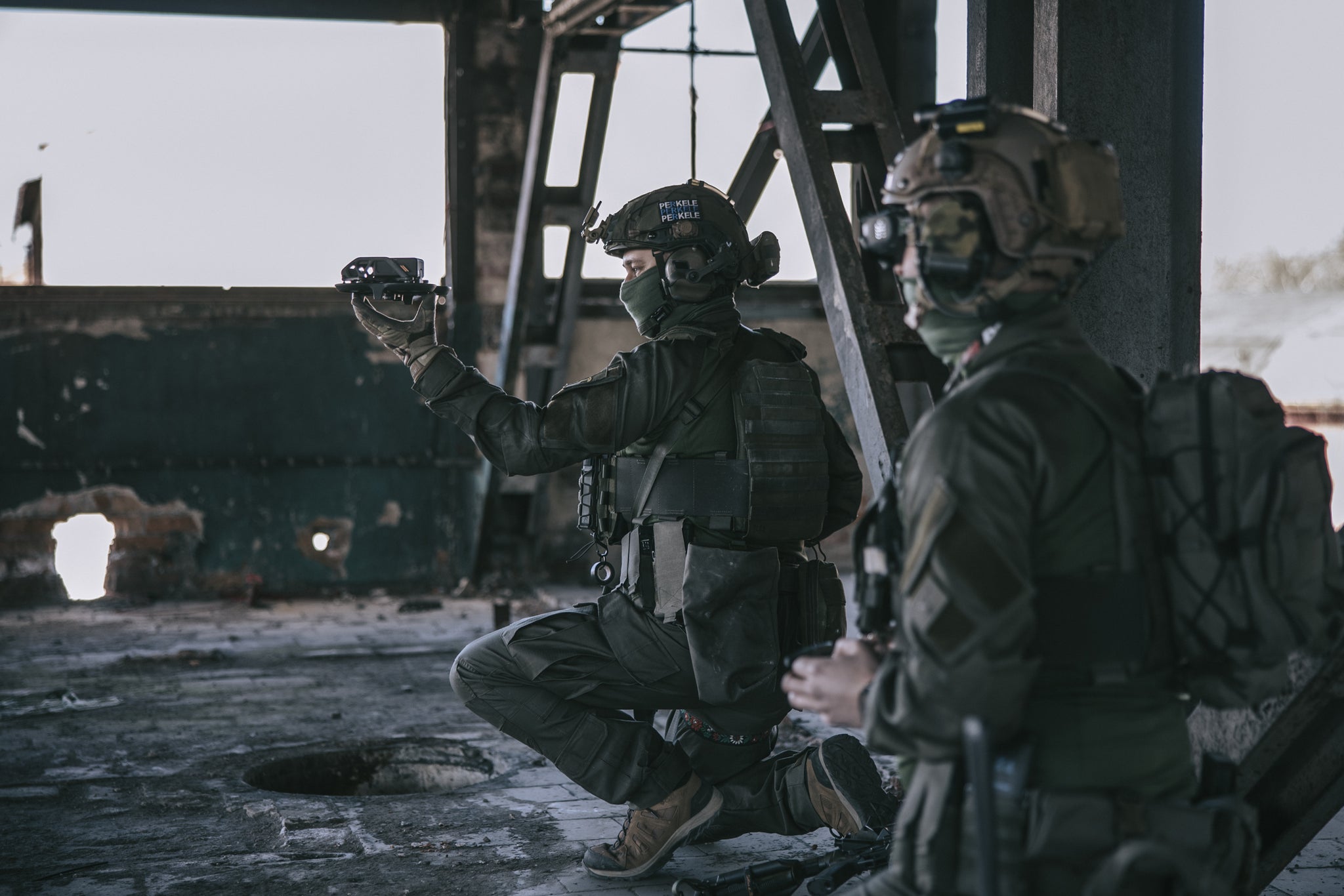 Two soldiers in tactical gear inside a dilapidated building, one of whom is holding a drone in his hands, ready to launch it.