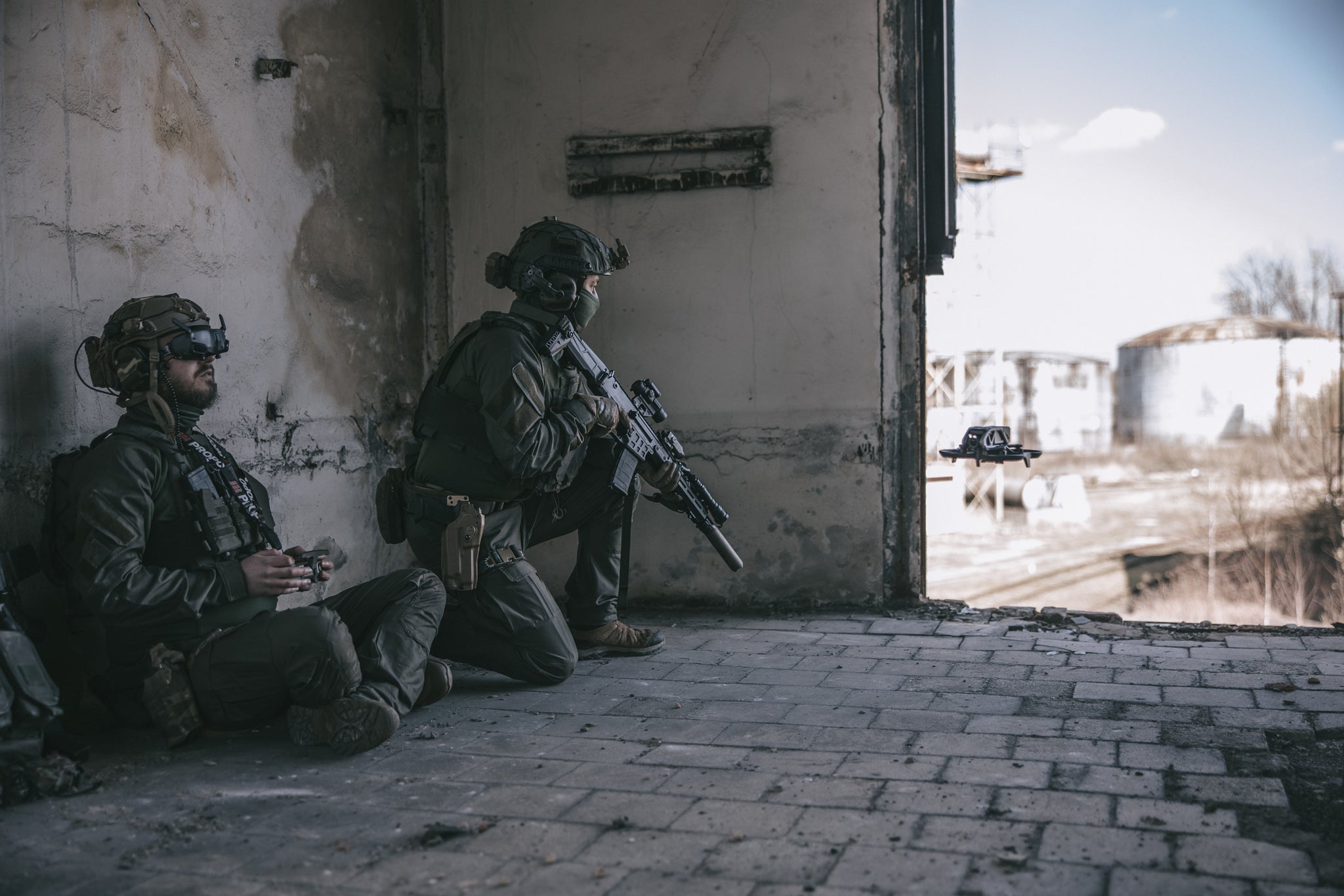 Two soldiers in tactical gear in an urban setting, one of whom is flying a drone, and the other is keeping a lookout with a rifle.