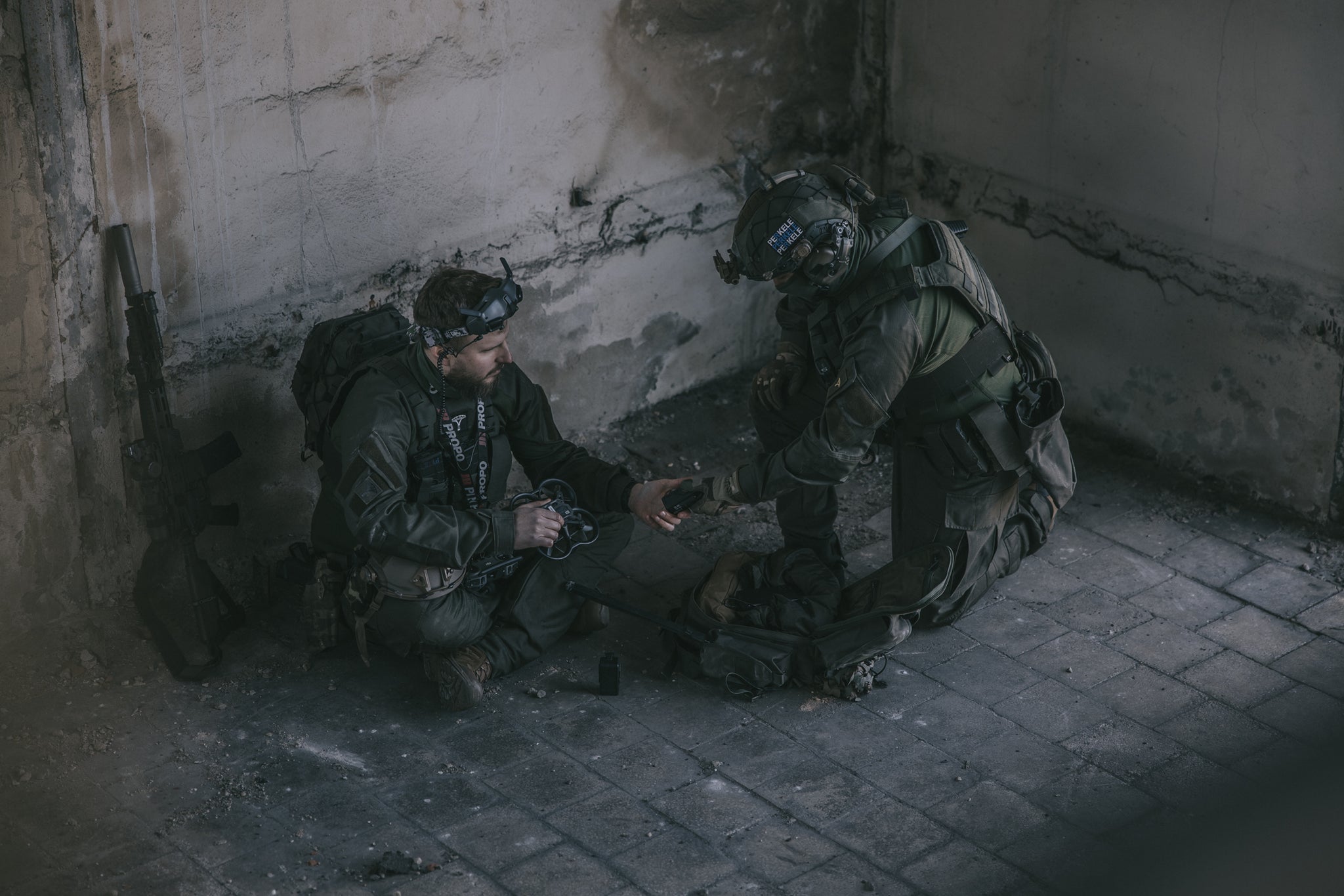 Two soldiers in tactical gear sitting on a concrete floor in a dimly lit room.