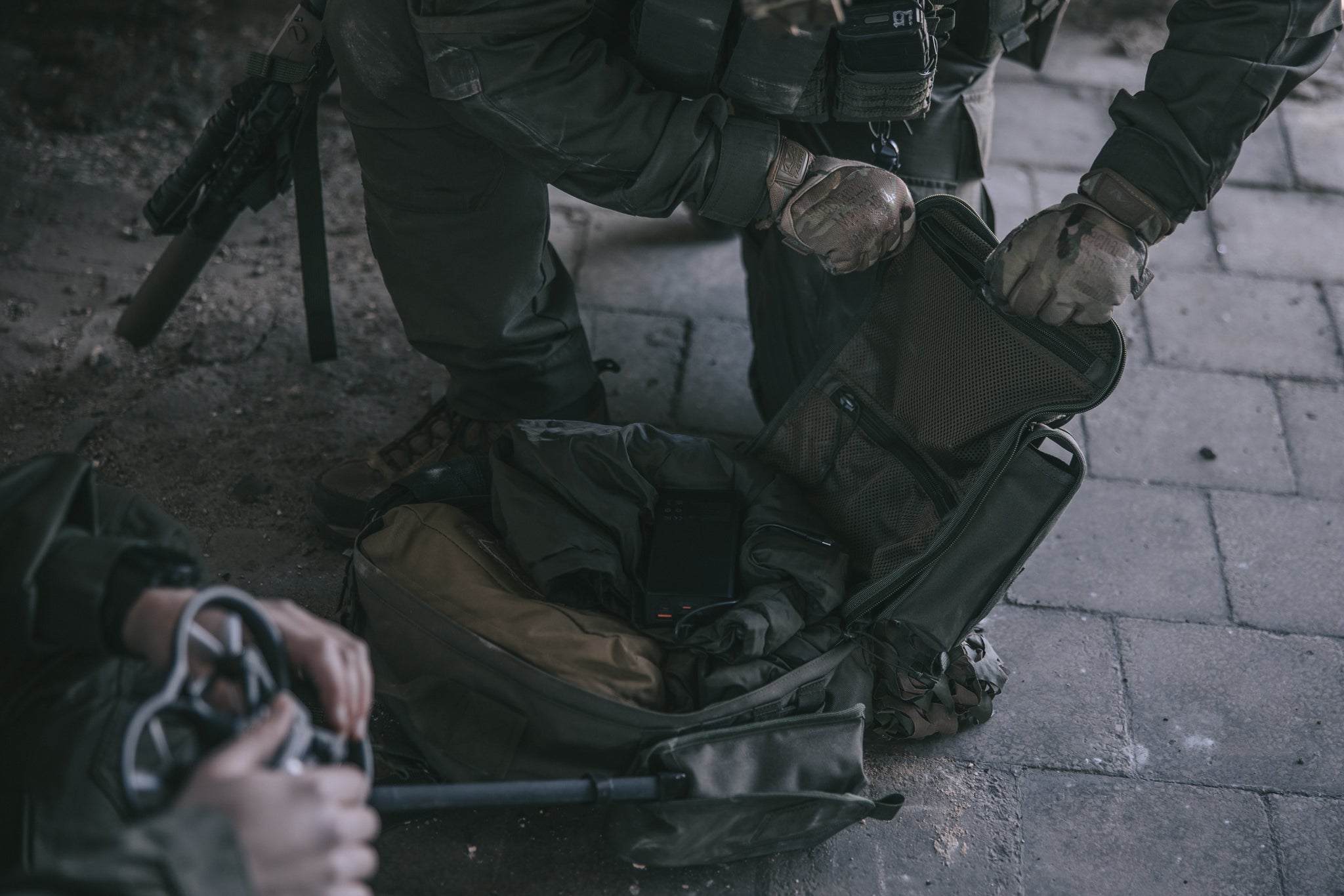 Person in tactical gear interacting with a backpack on a paved surface. You can see another person holding a drone in the foreground.