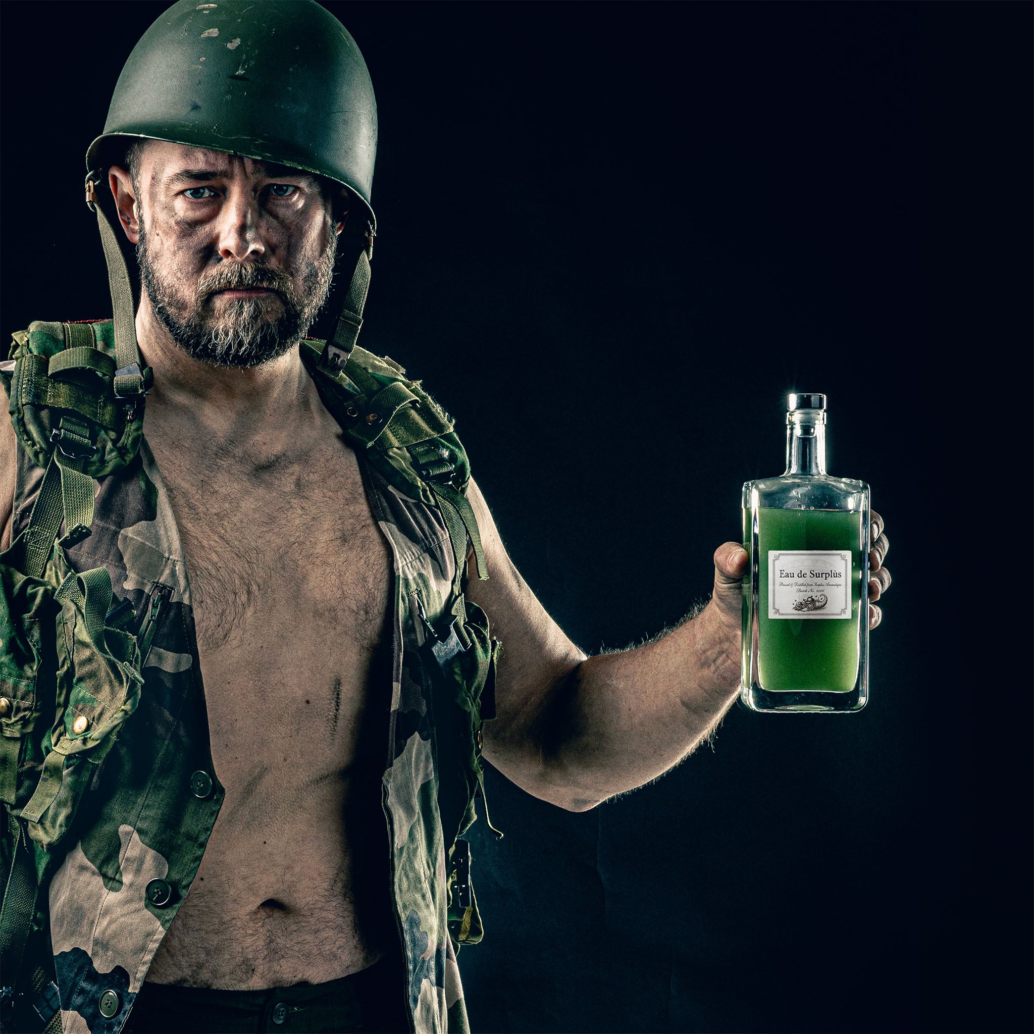 A man in an old military-style vest and helmet that is slightly scewed holding a bottle with green liquid in it on a dark background. The man has some dark marks on his face and arms, and the bottle is labelled Eau de Surplús.