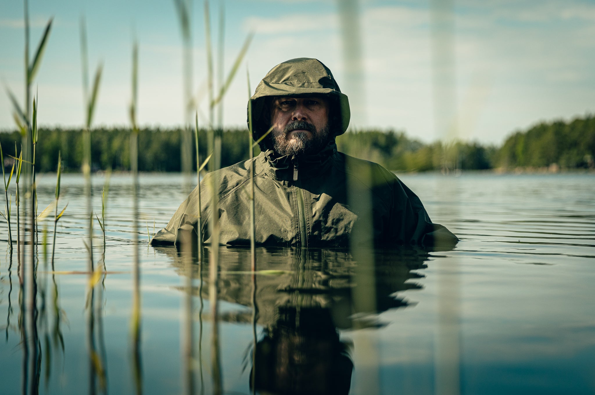 Man in a green jacket standing in water with tall grasses and trees in the background