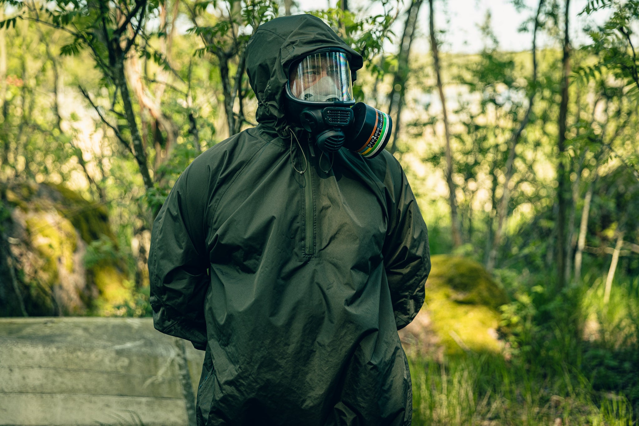 Person wearing a dark raincoat and a gasmask in a forest setting