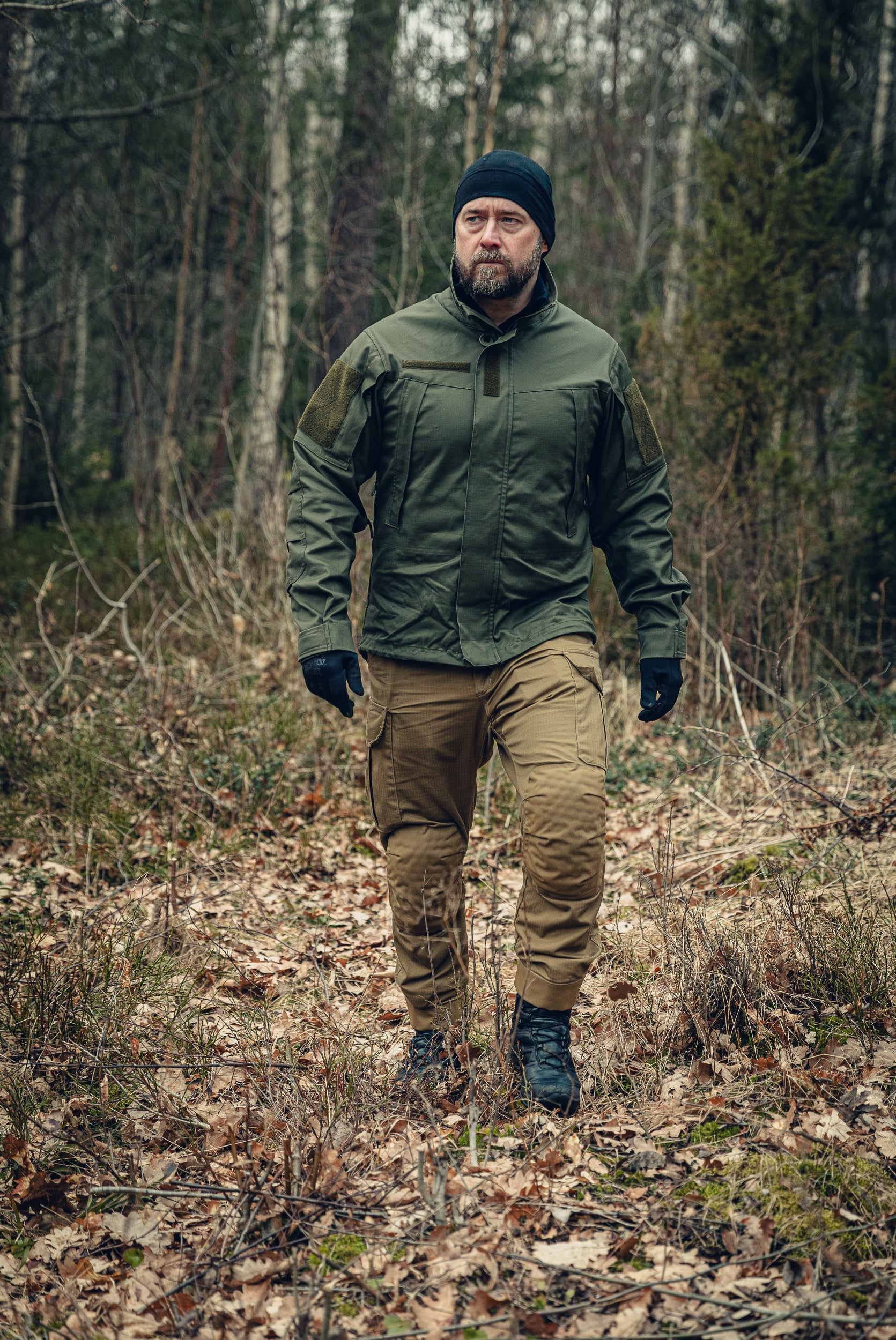 A bearded man walking in a forest looking slightly to the left. He is wearing a green jacket with a snug fit, a black beanie and gloves and light brown cargo pants.