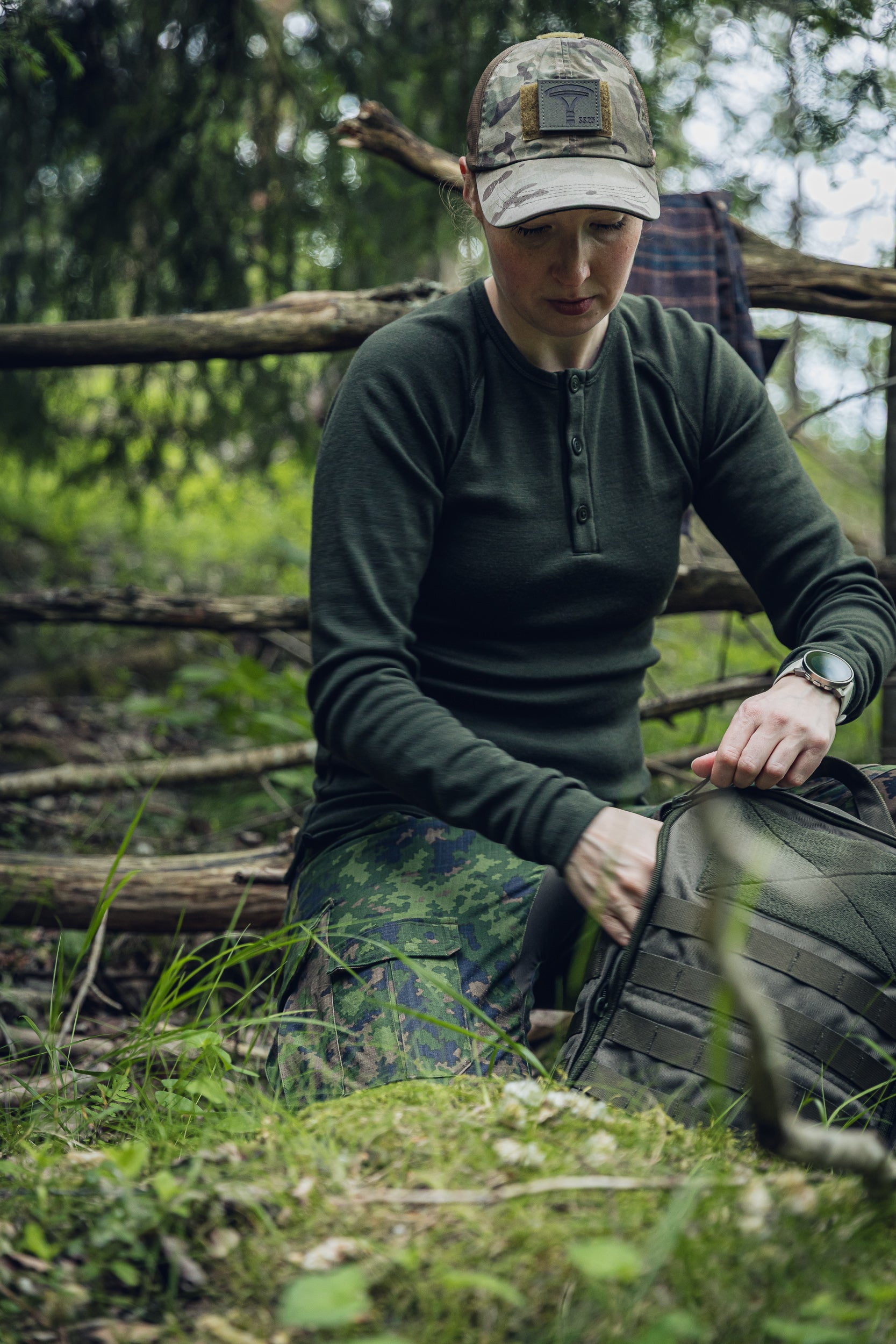 A woman kneeling outdoors in a wooded setting, wearing a dark olive long-sleeve henley shirt, camouflage pants, and a camouflage cap while packing or organizing a dark tactical-style bag.