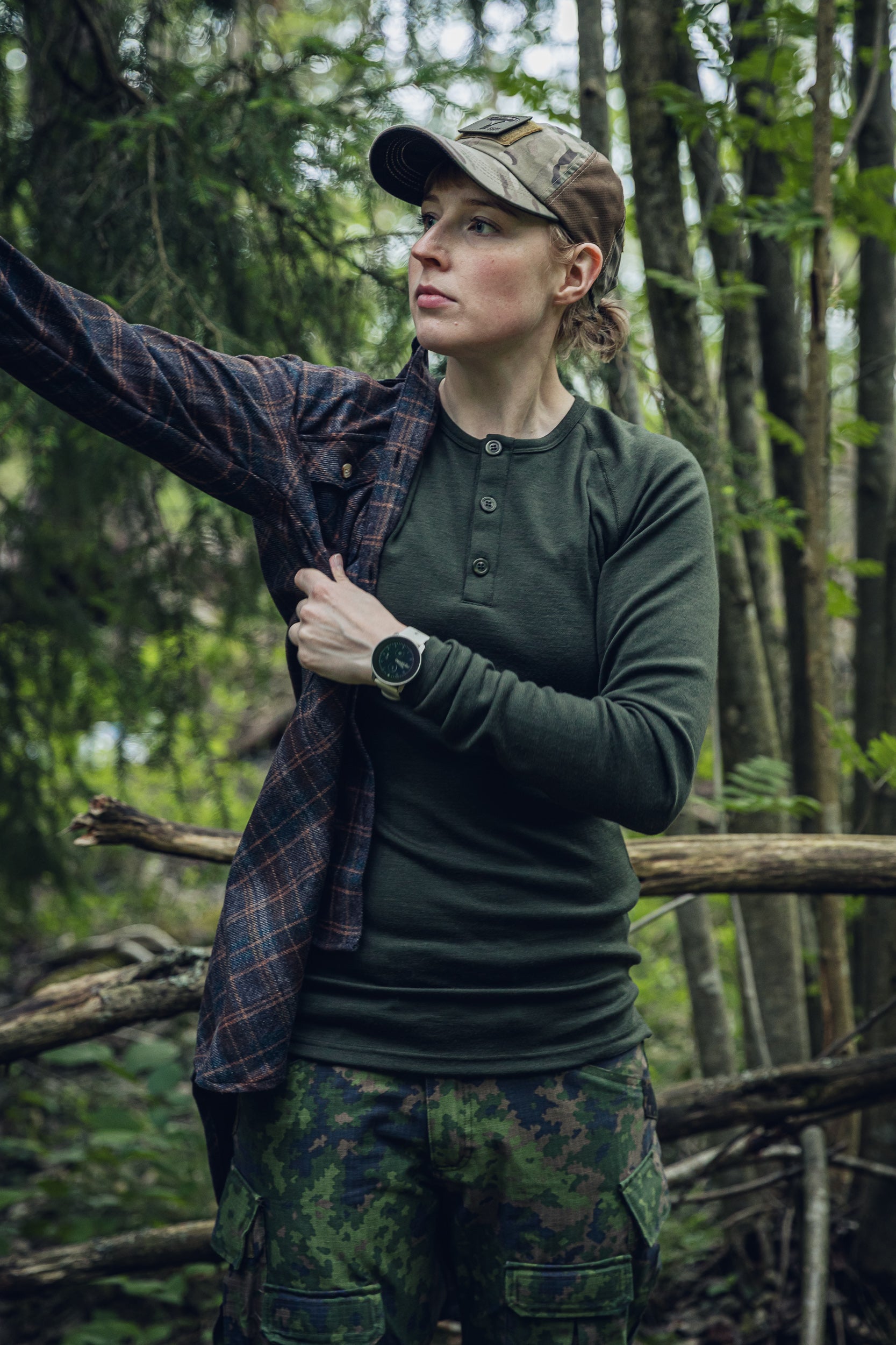 A woman in a forest wearing a dark olive green long-sleeve henley shirt, camouflage pants, a camouflage cap, and a dark plaid overshirt.