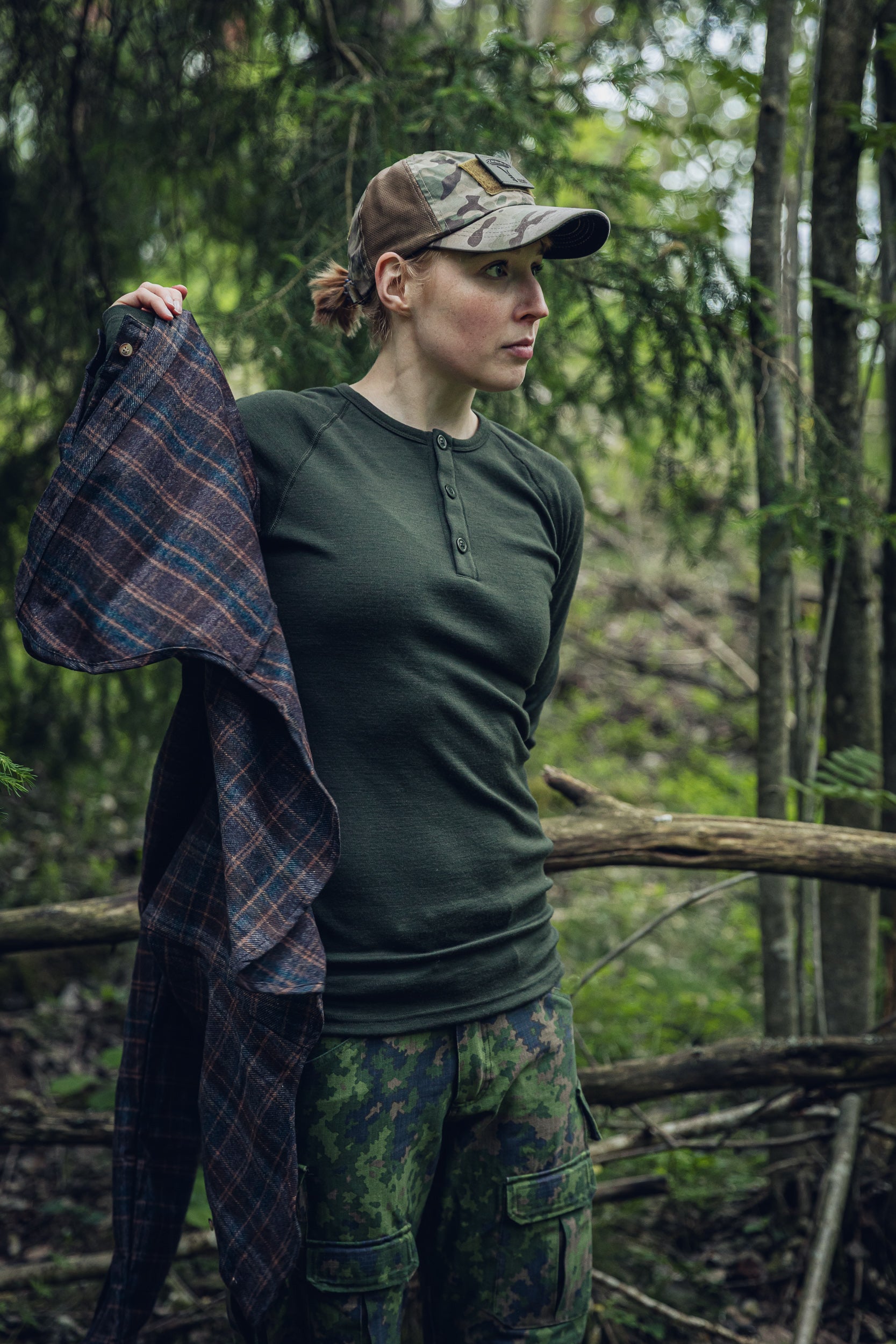 A woman standing in a forest wearing a dark olive green Henley shirt, camouflage pants, and a camouflage cap, holding a plaid flannel shirt over her shoulder.