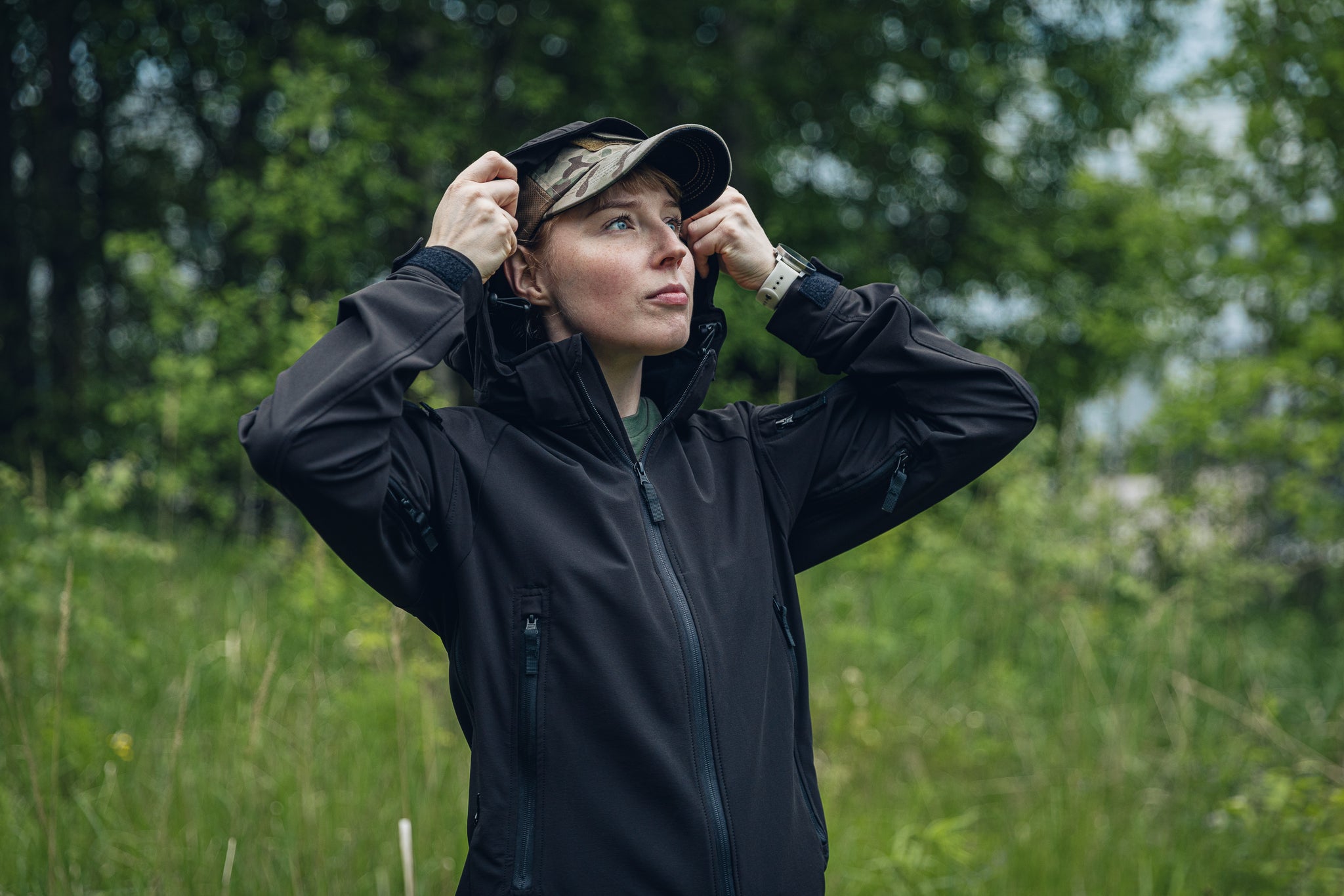 A woman wearing a black outdoor  jacket and a camouflage cap stands in a grassy field with trees in the background, adjusting her hood.