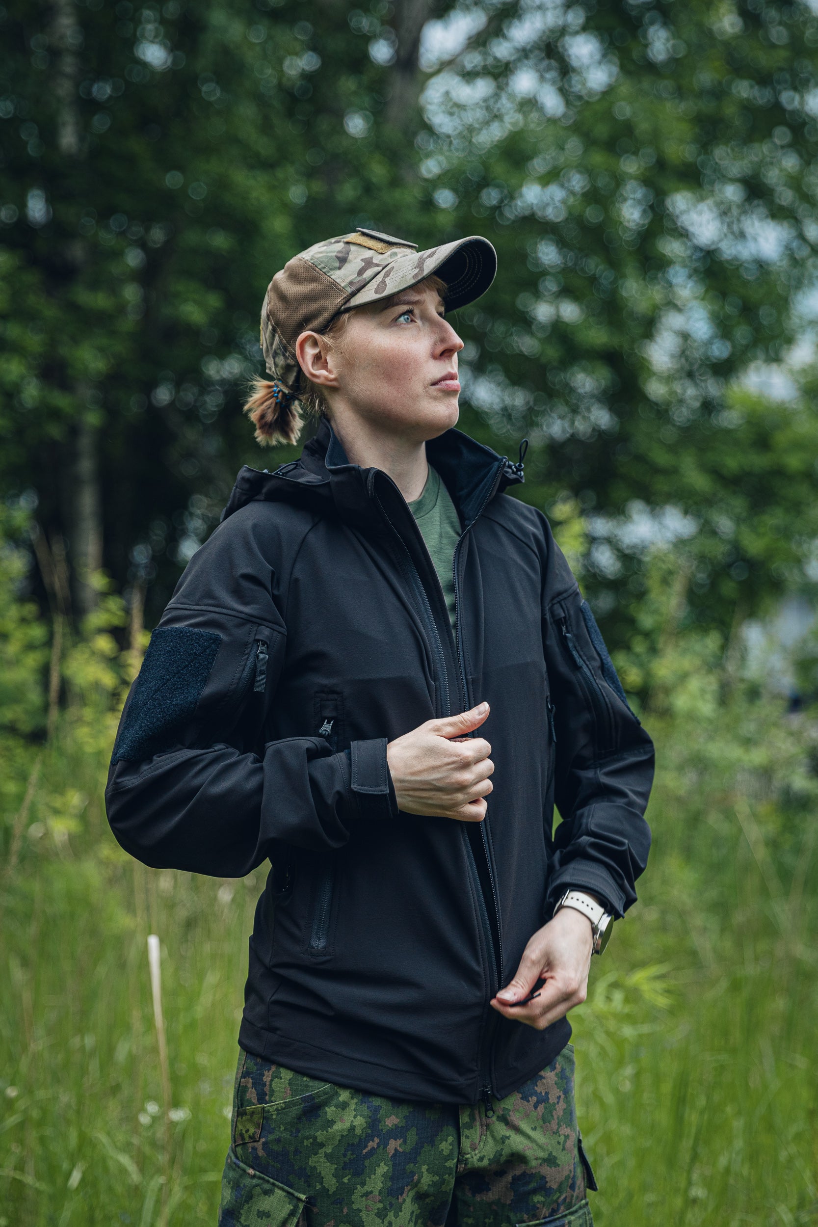 A woman wearing a black tactical-style jacket, camouflage cap, and camouflage pants stands outdoors in a grassy wooded area, looking to the side.