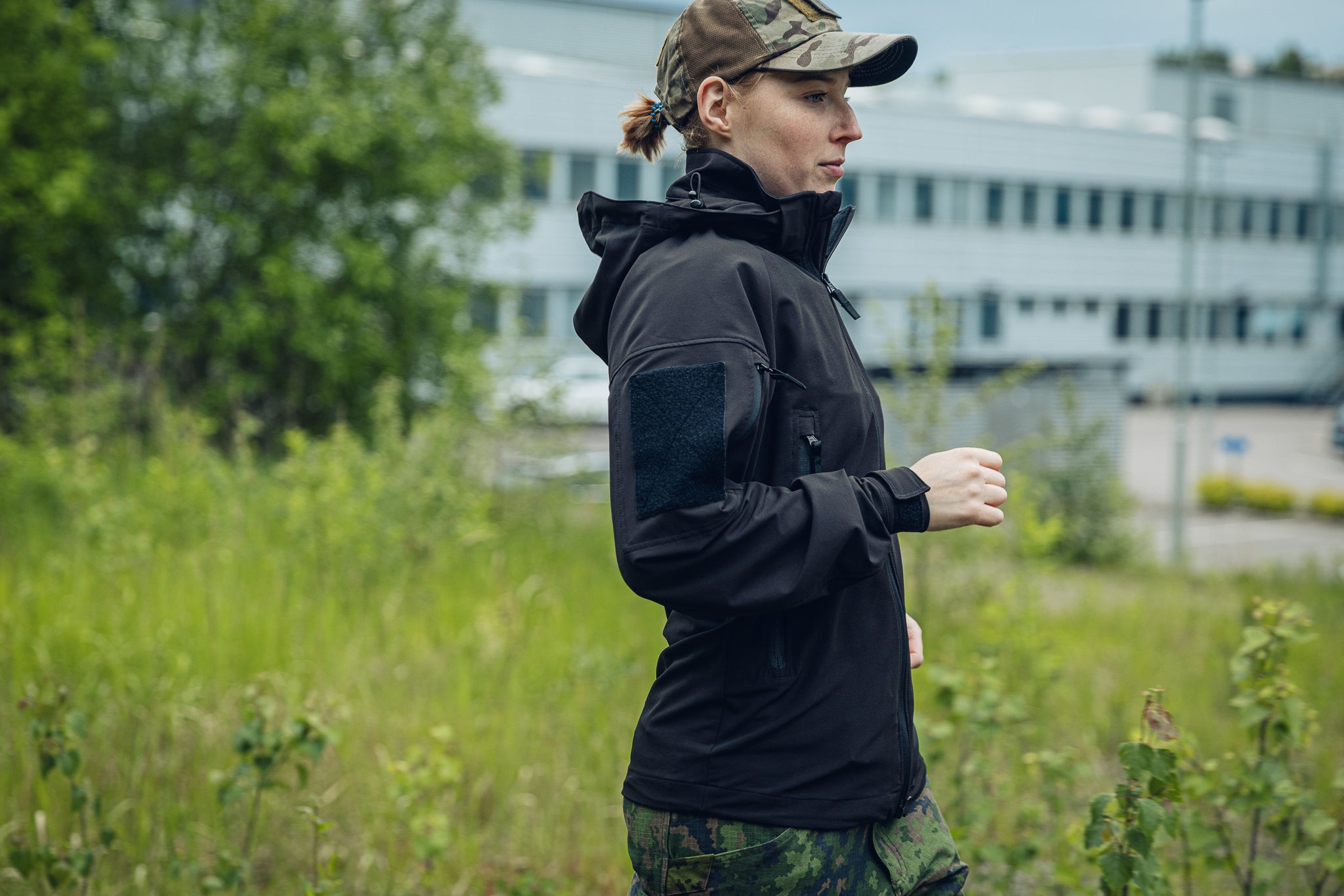 A woman wearing a black hooded tactical jacket and camouflage pants stands outdoors in a grassy area with a building in the background.