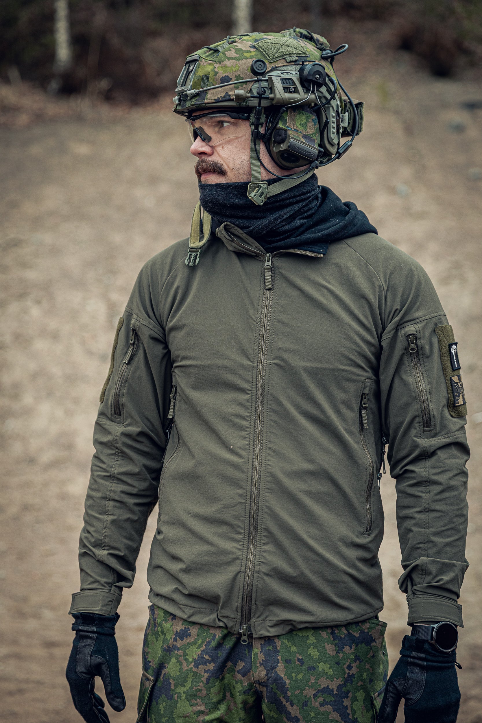 Person wearing military gear including a helmet, jacket, and gloves in an outdoor setting.