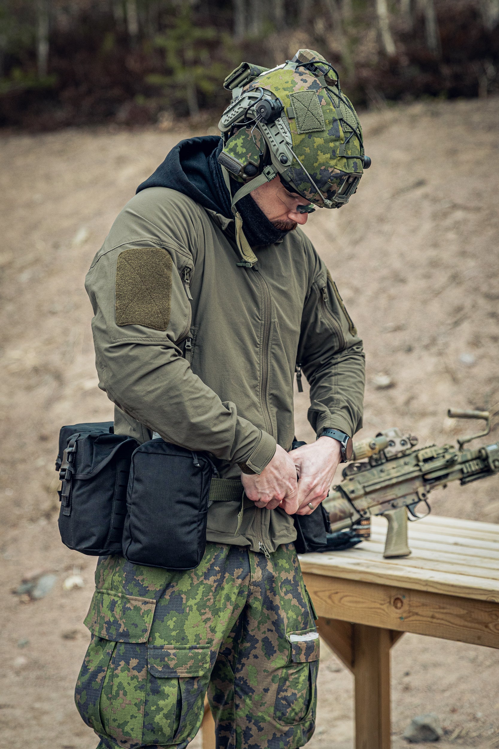 A man in military-style camouflage clothing and a helmet stands outdoors at a wooden table, handling a rifle. The scene includes a real outdoor environment with dirt ground and trees in the background.