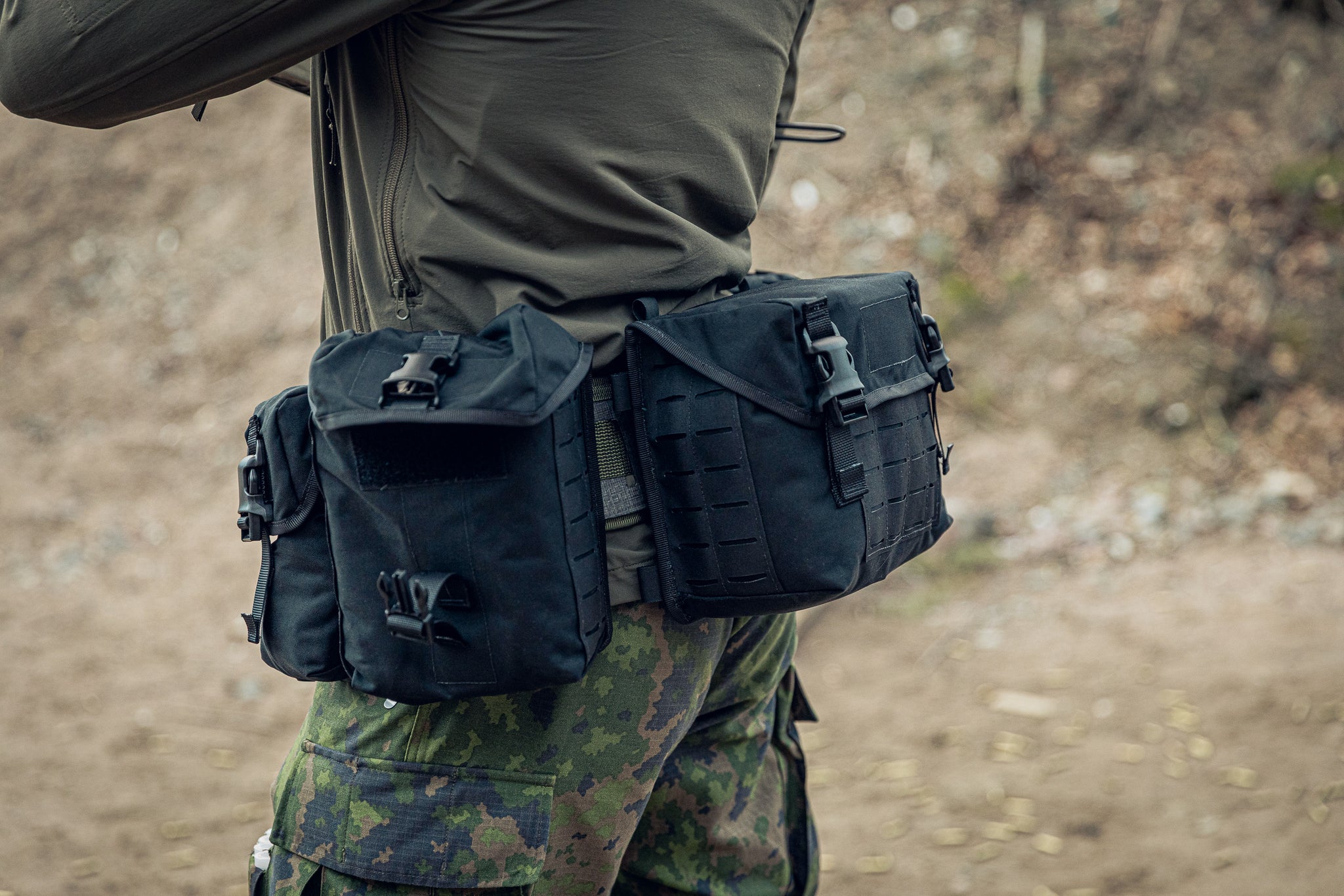 A close-up of a person outdoors wearing tactical gear, including a black waist pouch system with three black general purpose pouches and camouflage pants.