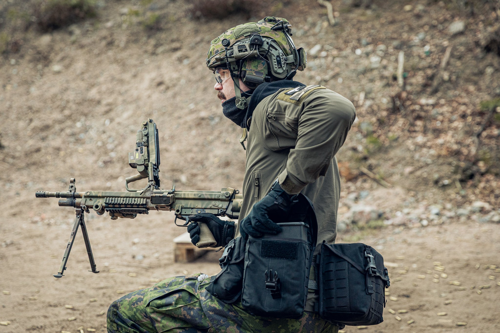 A person in military-style camouflage clothing and helmet kneels on a dirt slope while holding a mounted rifle. The scene is outdoors with a blurred natural background.