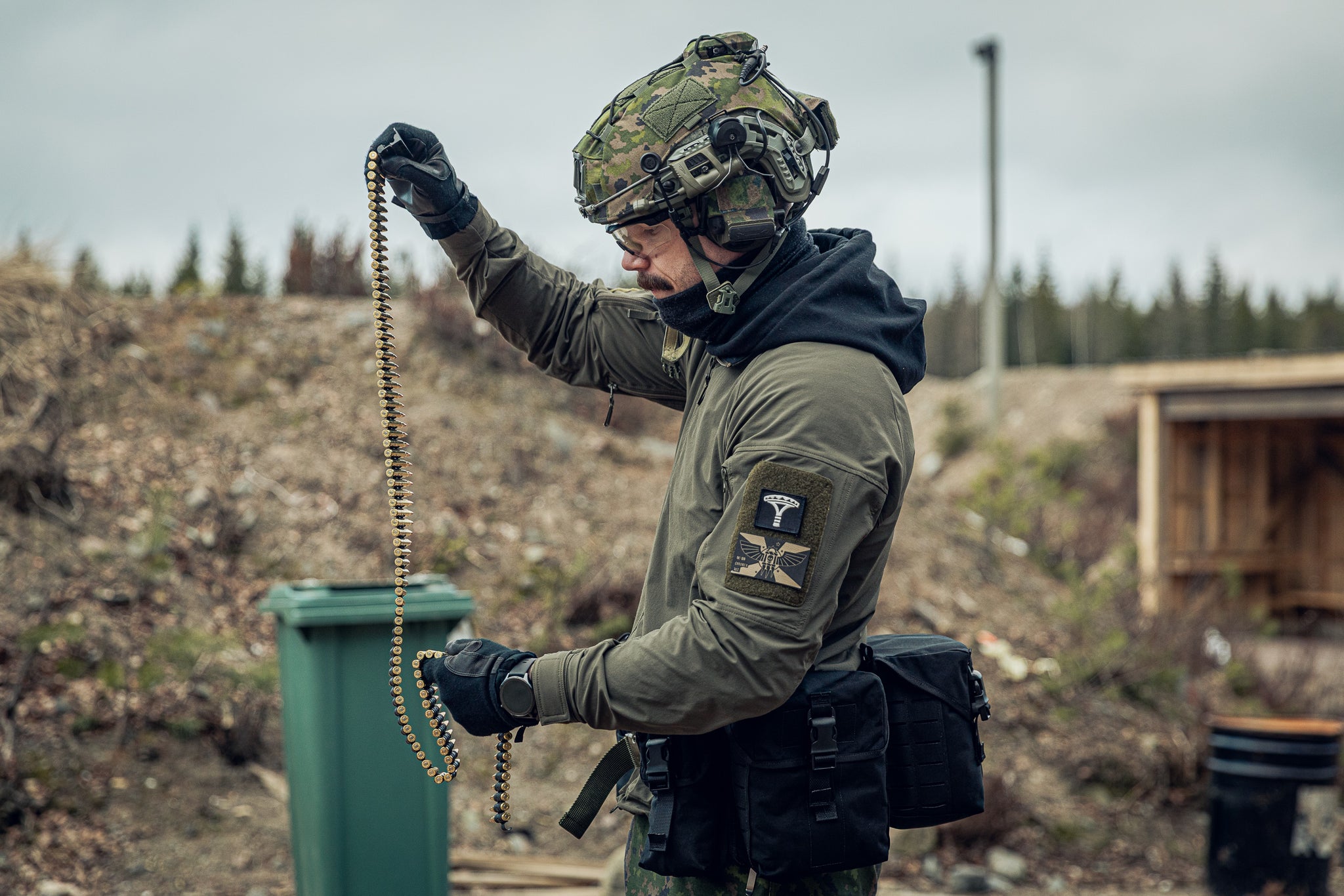 A person in military-style gear, including a camouflage helmet and an olive drab jacket, stands outdoors, holding a chain of ammunition. The background shows a barren landscape, a green trash can, and a small wooden structure under an overcast sky.