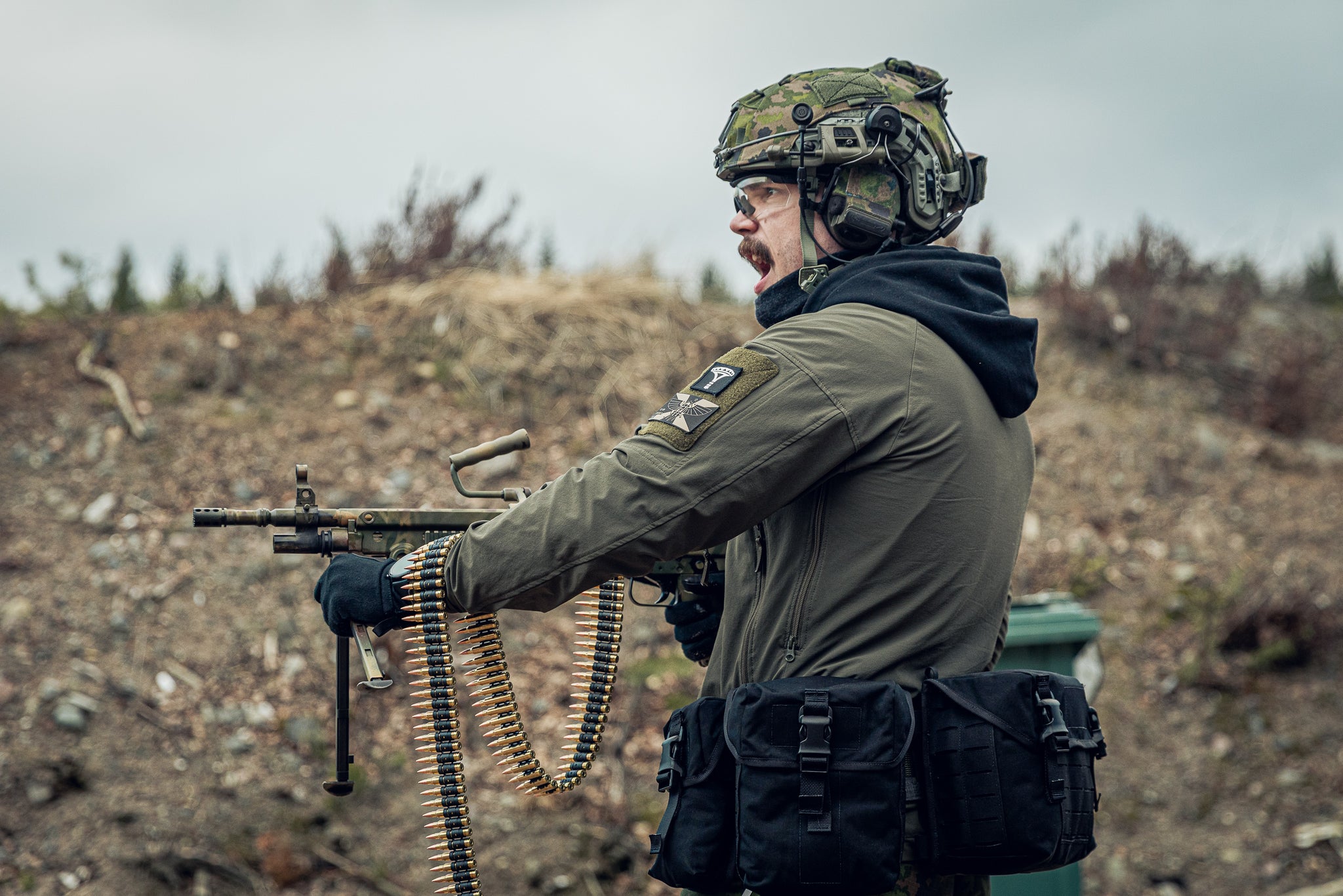 A person in tactical gear, including a camouflage helmet, olive drab jacket, and black utility pouches, holding a submachine gun with a mounted belt loader in an outdoor, rugged terrain setting.