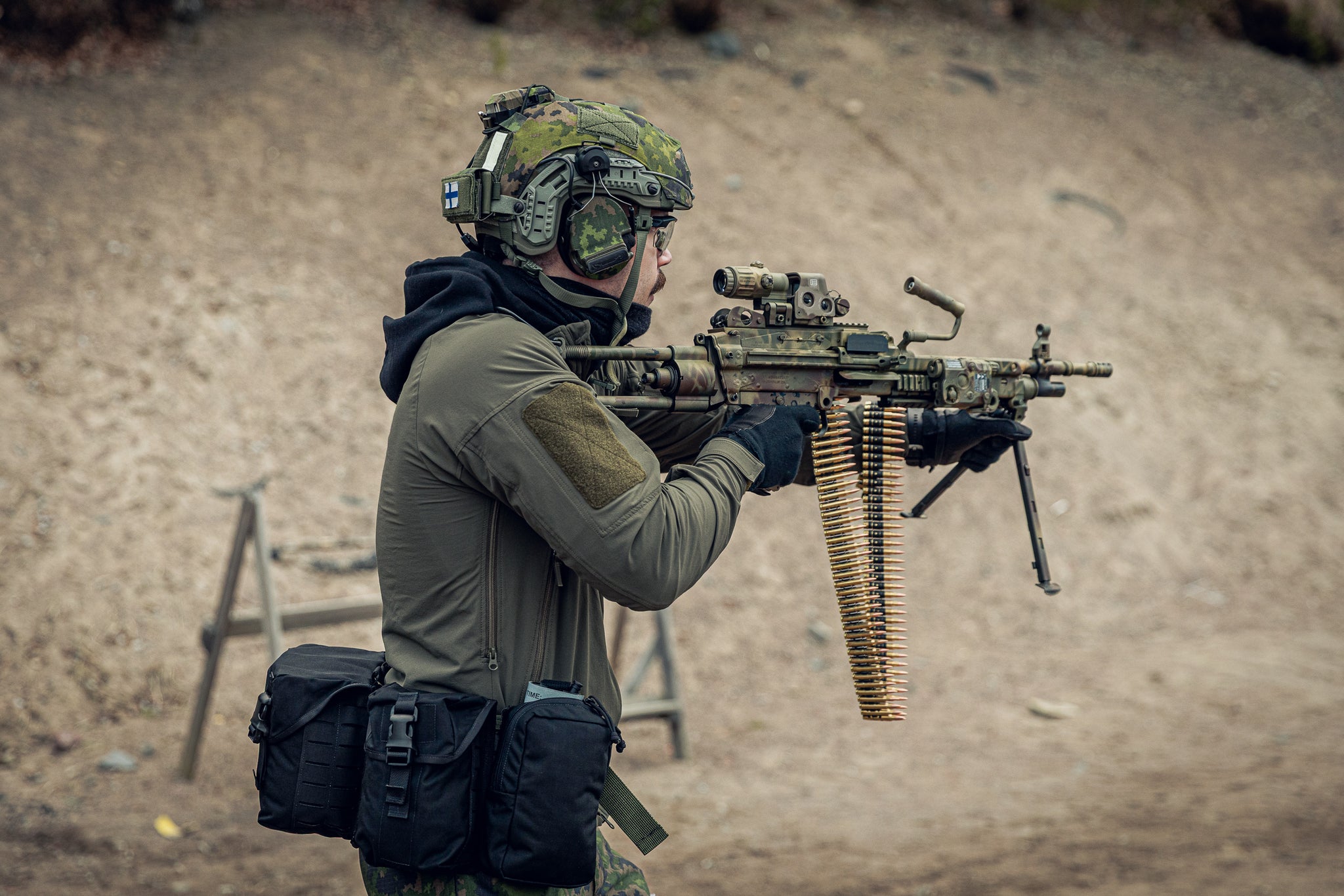 A person in tactical gear, including a camouflage helmet, ear protection, and a green hooded jacket, aiming a mounted machine gun with a dangling ammunition belt in an outdoor sandy setting.