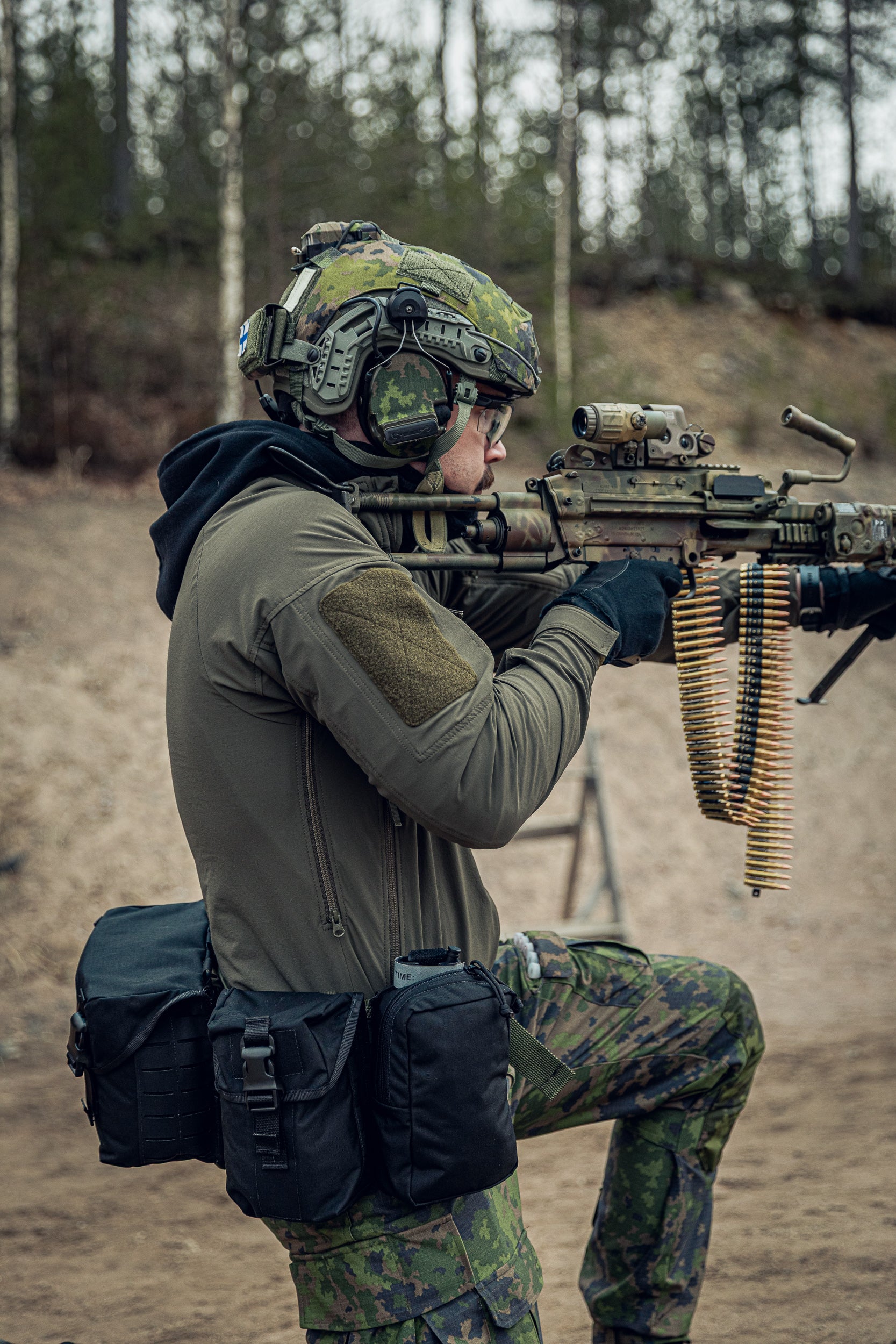 A person in tactical military-style clothing and a camouflage helmet aims a rifle outdoors in a wooded, sandy terrain. The outfit includes an olive drab jacket, camouflage pants, black gloves, and a black utility pouch at the hip.