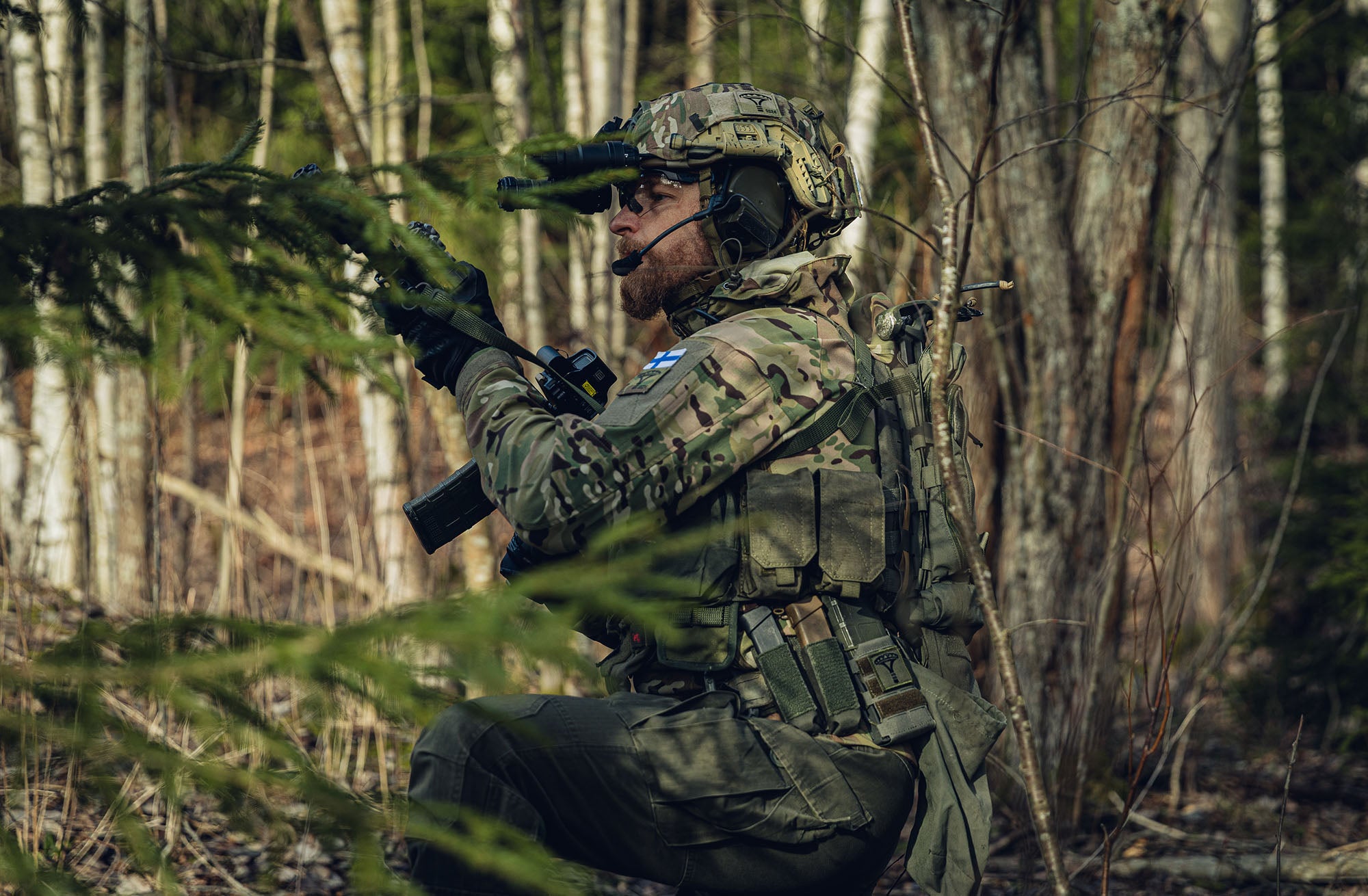 A soldier kneeling in a forest with a rifle in his hand. He is wearing a helmet with  night-vision goggles and hearing protection attached to them, a multicam jacket, a plate carrier and green cargo pants.