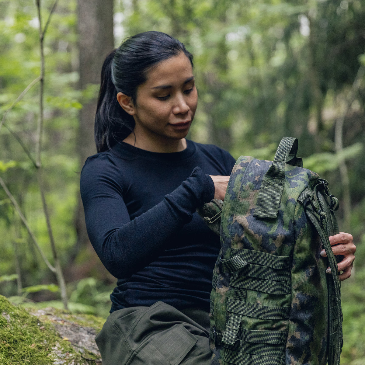 A woman in a black long-sleeve shirt and olive pants packs items into a green camouflage tactical backpack in a forest setting.
