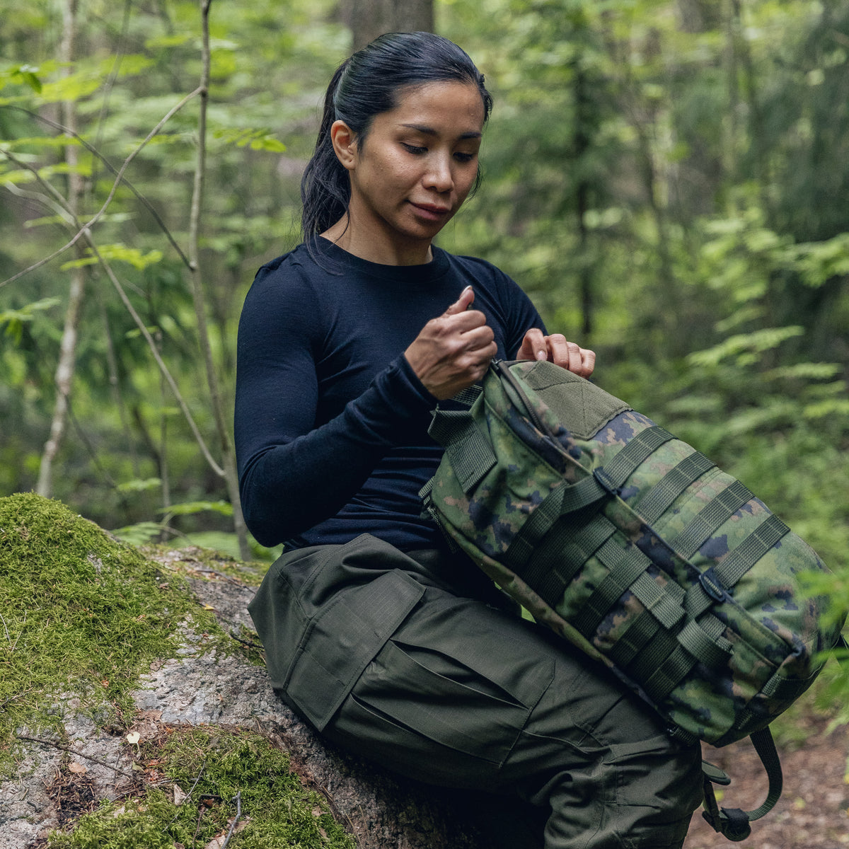 A woman in a forest setting sits on a mossy log, organizing gear in a green camouflage tactical backpack.