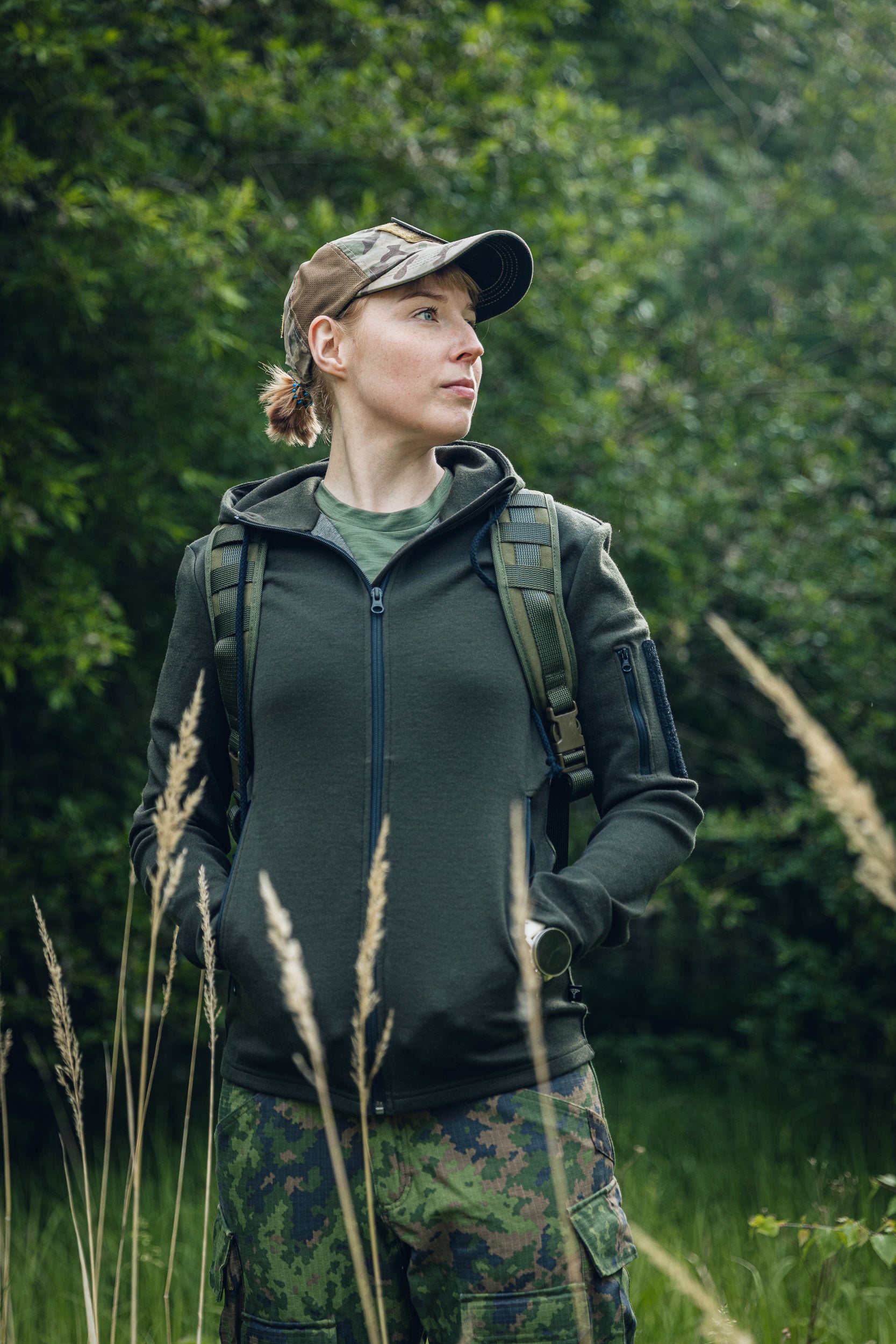 A woman wearing a dark olive green zip-up hoodie, camouflage pants, and a matching cap stands in a grassy, wooded outdoor setting.