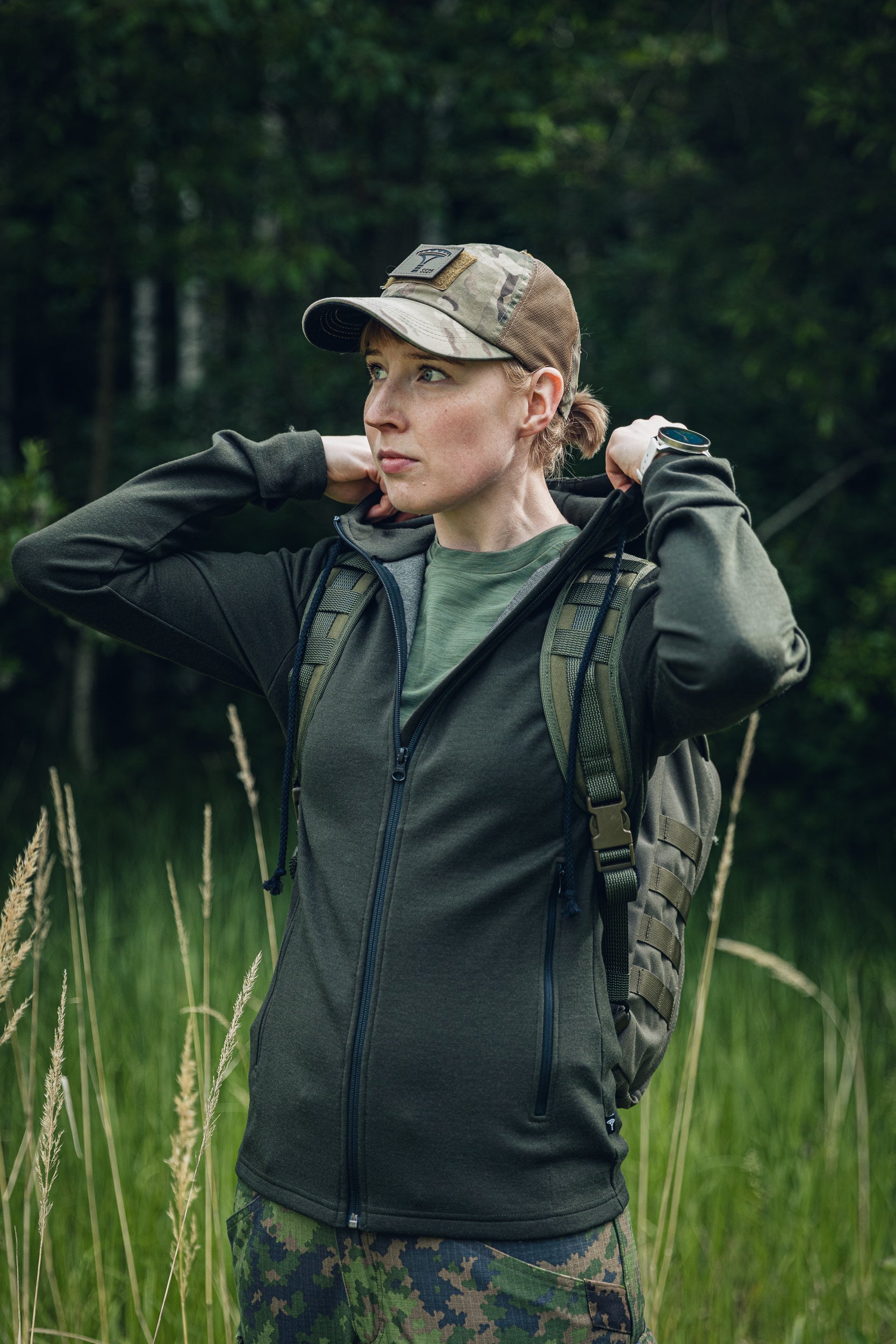 A woman outdoors in a grassy field wearing a dark olive green zip-up hoodie, a camouflage cap, and a tactical backpack.