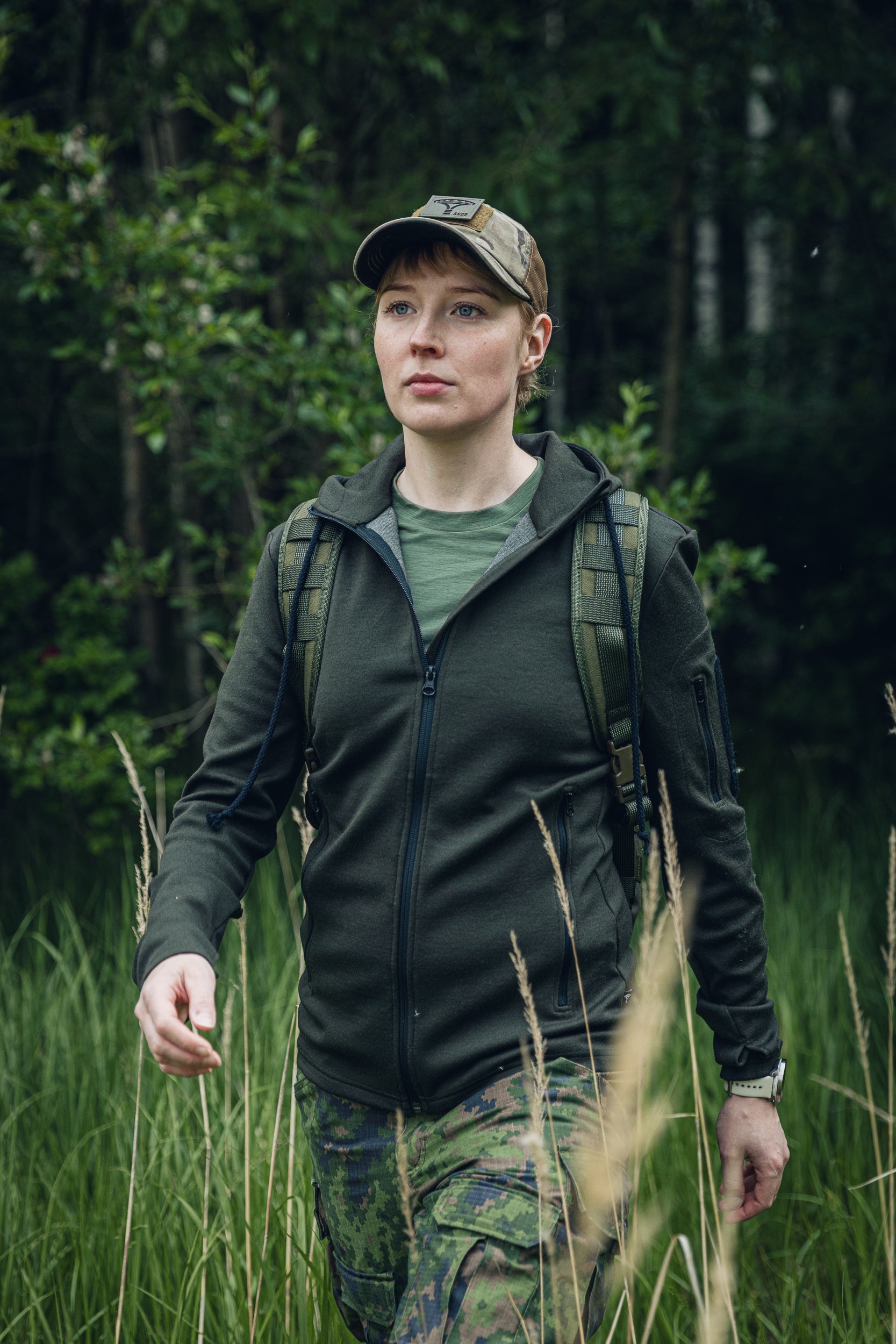 A woman wearing a dark olive green zip-up hoodie, camouflage pants, and a matching camouflage cap walks through tall grass in a forest setting.