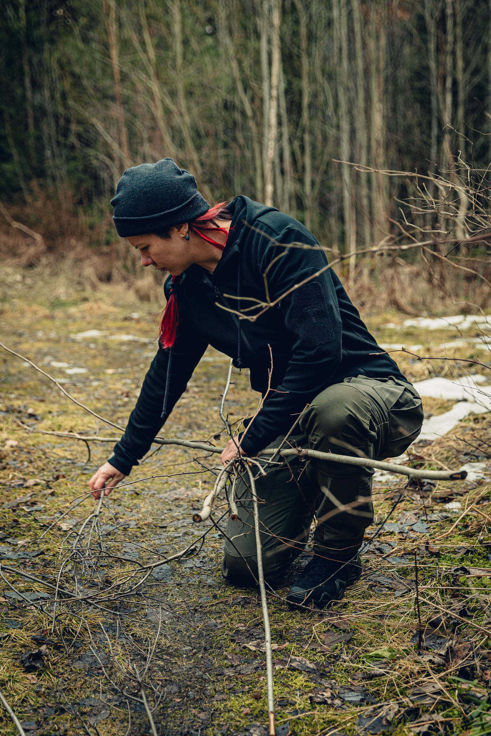 A woman kneeling on the ground gathering some sticks. She is wearing green cargo pants, a black hoodie and a grey beanie.