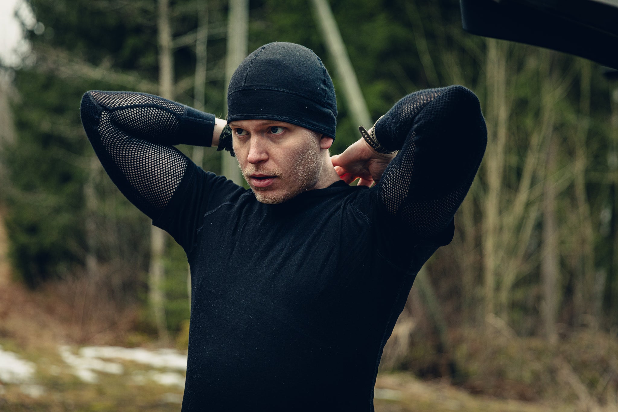 A young blonde man wearing a black t-shirt, black beanie and black long-sleeved mesh shirt. He seems to be adjusting the back of his shirt.There is a slightly snowy forest in the background.