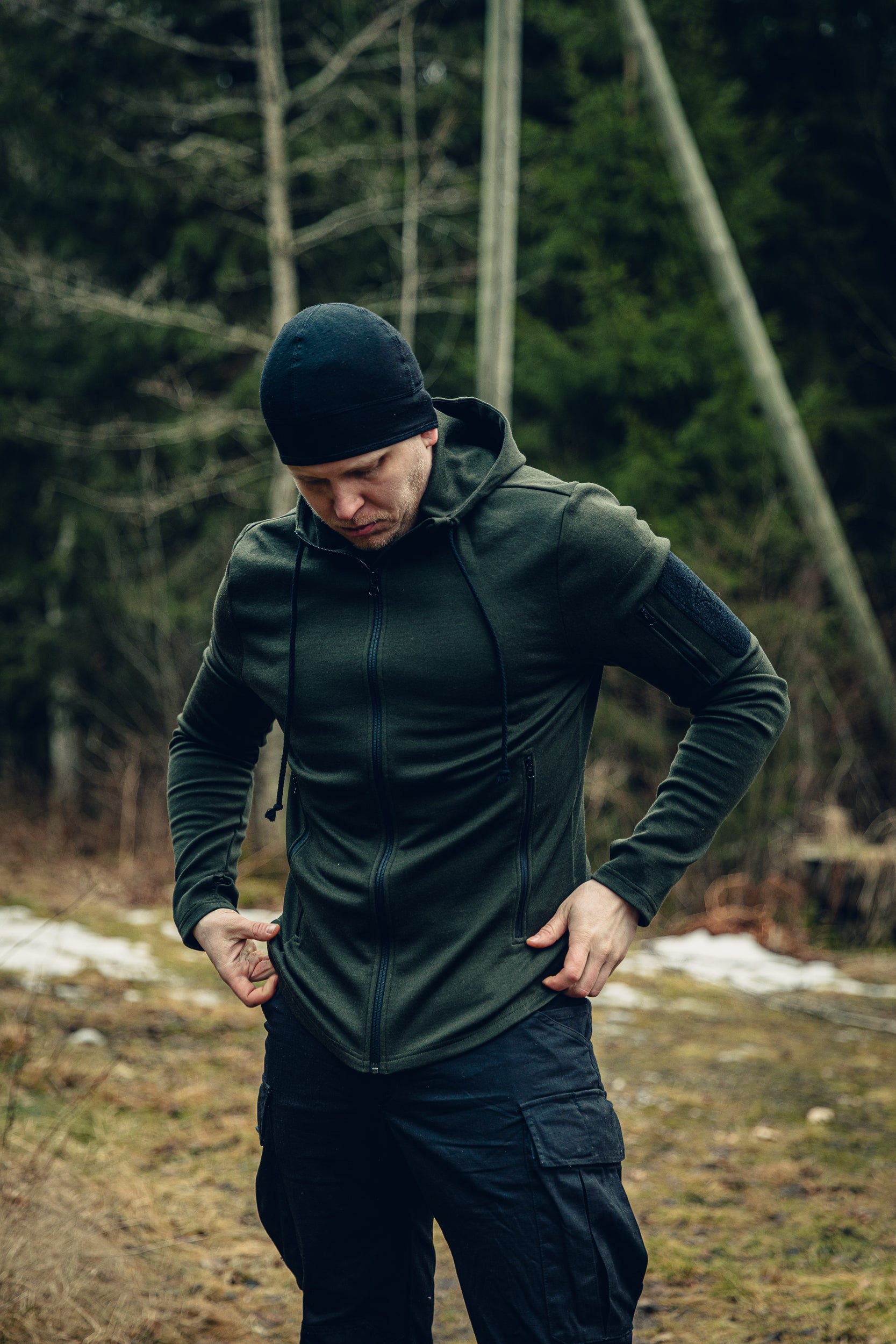 A man standing in a yard with a forest in the background, adjusting the hem of his green hoodie. He is also wearing a balck beanie and black cargo pants.