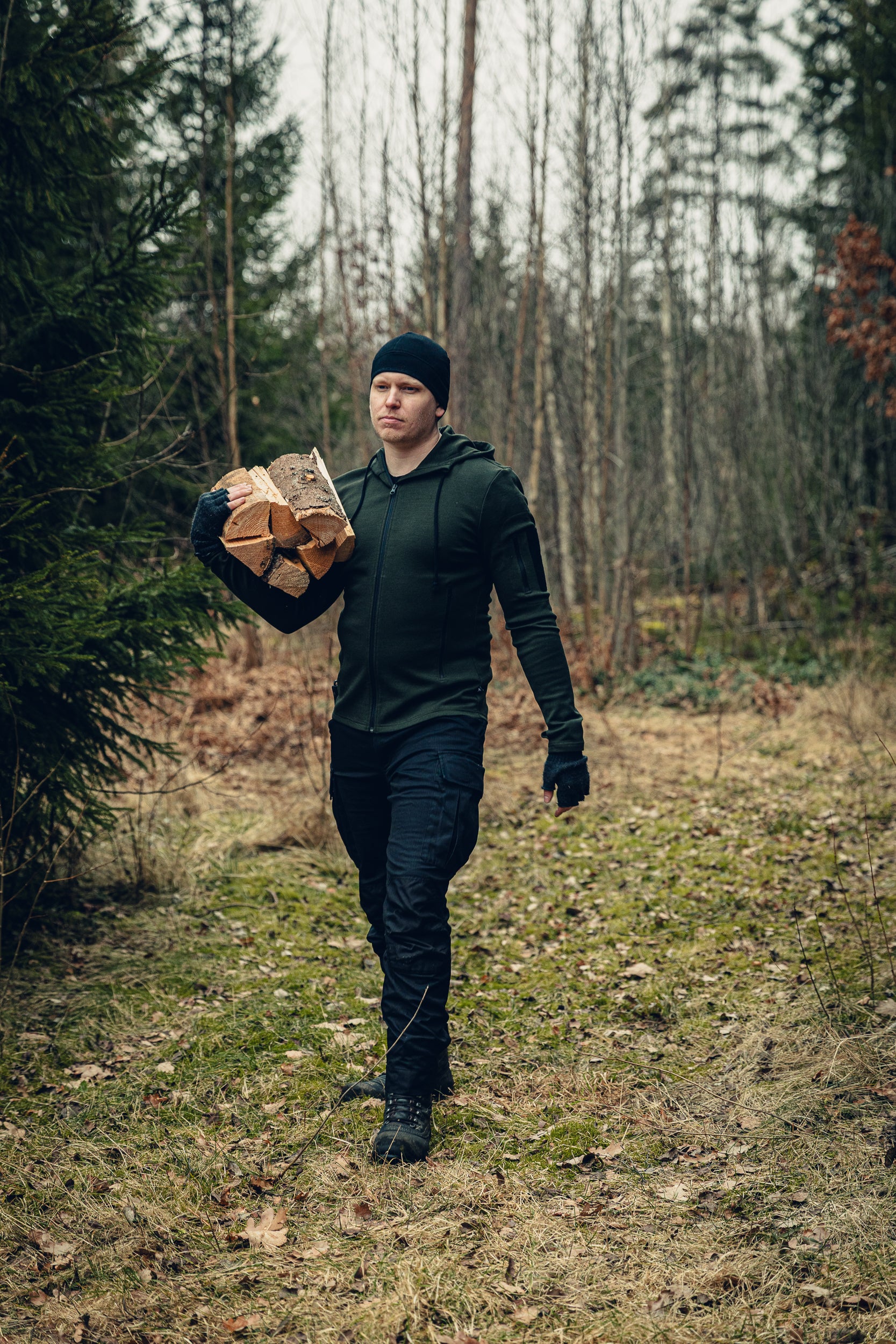 A man walking through a yard holding pieces of firewood. He is wearing a black beanie, green hoodie, fingerless gloves and black cargo pants. There is a forest in the background.