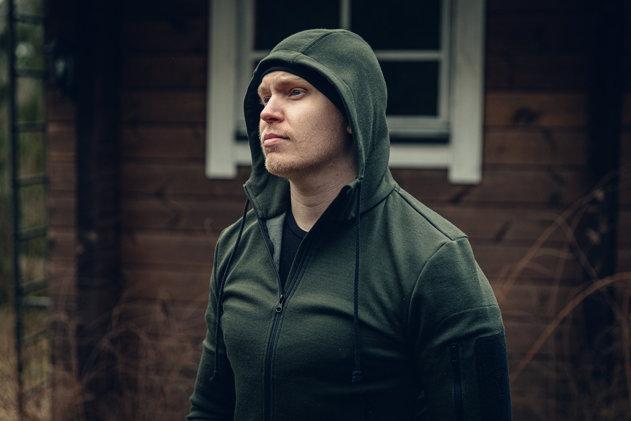 A man in front of an old wooden cabin, wearing a green hoodie with the hood pulled up.