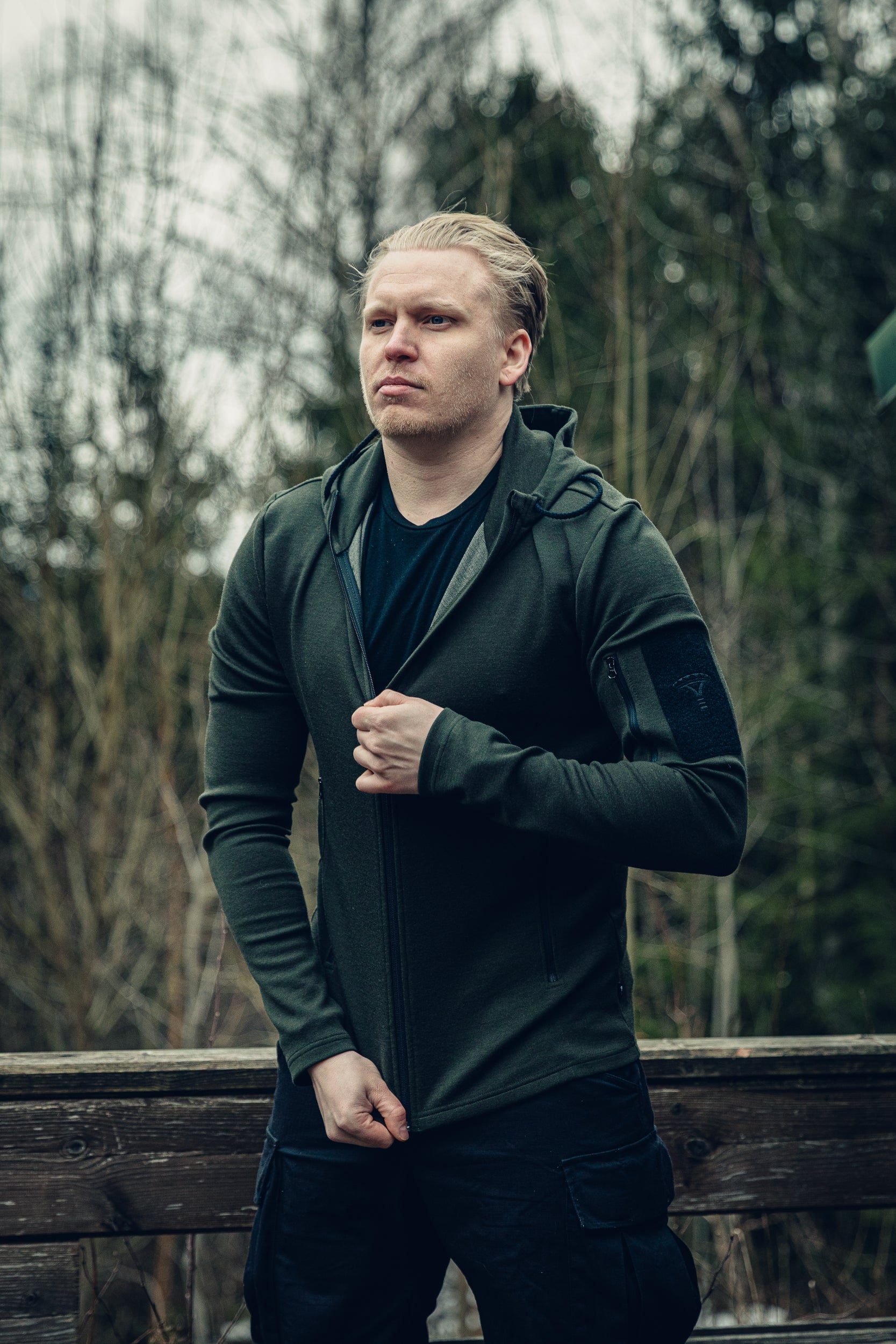 A man outside zipping up a dark green hoodie. There is a forest and a small wooden fence in the background. 