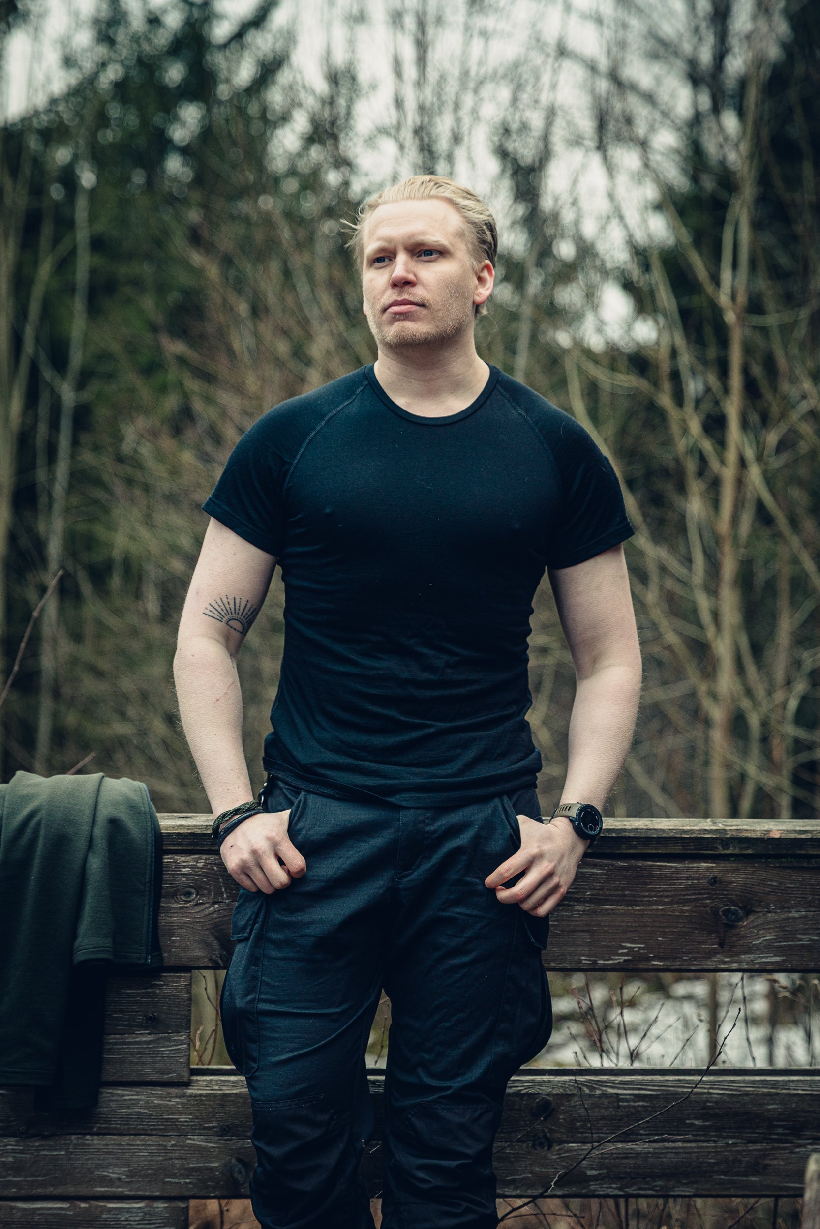A young blonde man casually leaning against a small wooden fence wearing a black t-shirt and black pants. There is a green garment next to him on the fence, and a slightly snowy forest in the background.
