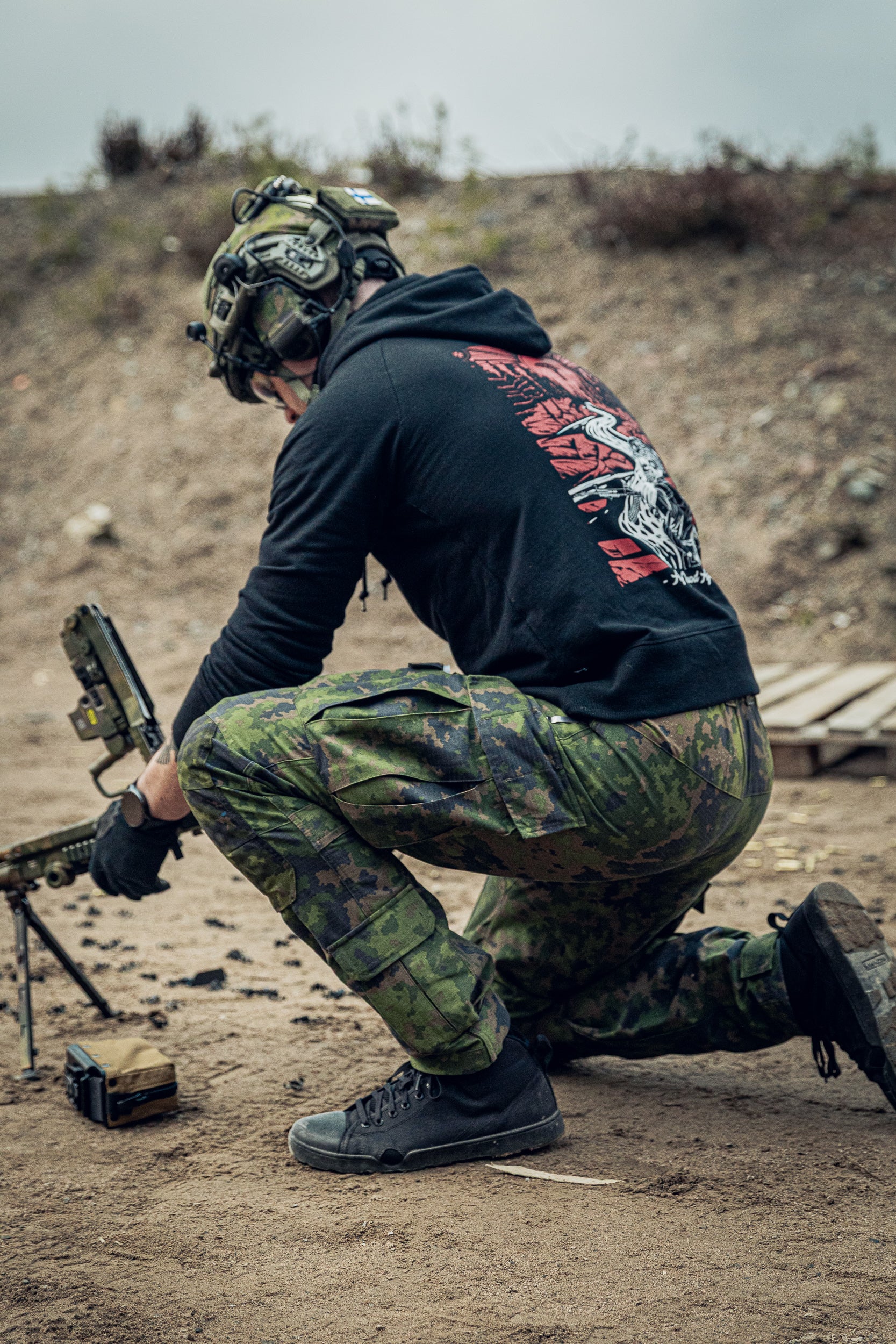 Person in camouflage gear and black hoodie with graphic design, kneeling on a dirt field.