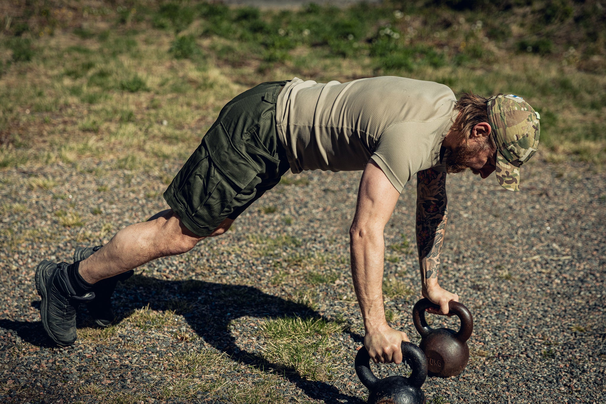 A man in a khaki t-shirt, multicam cap and green shorts doing pushups on top of two kettlebells outside.