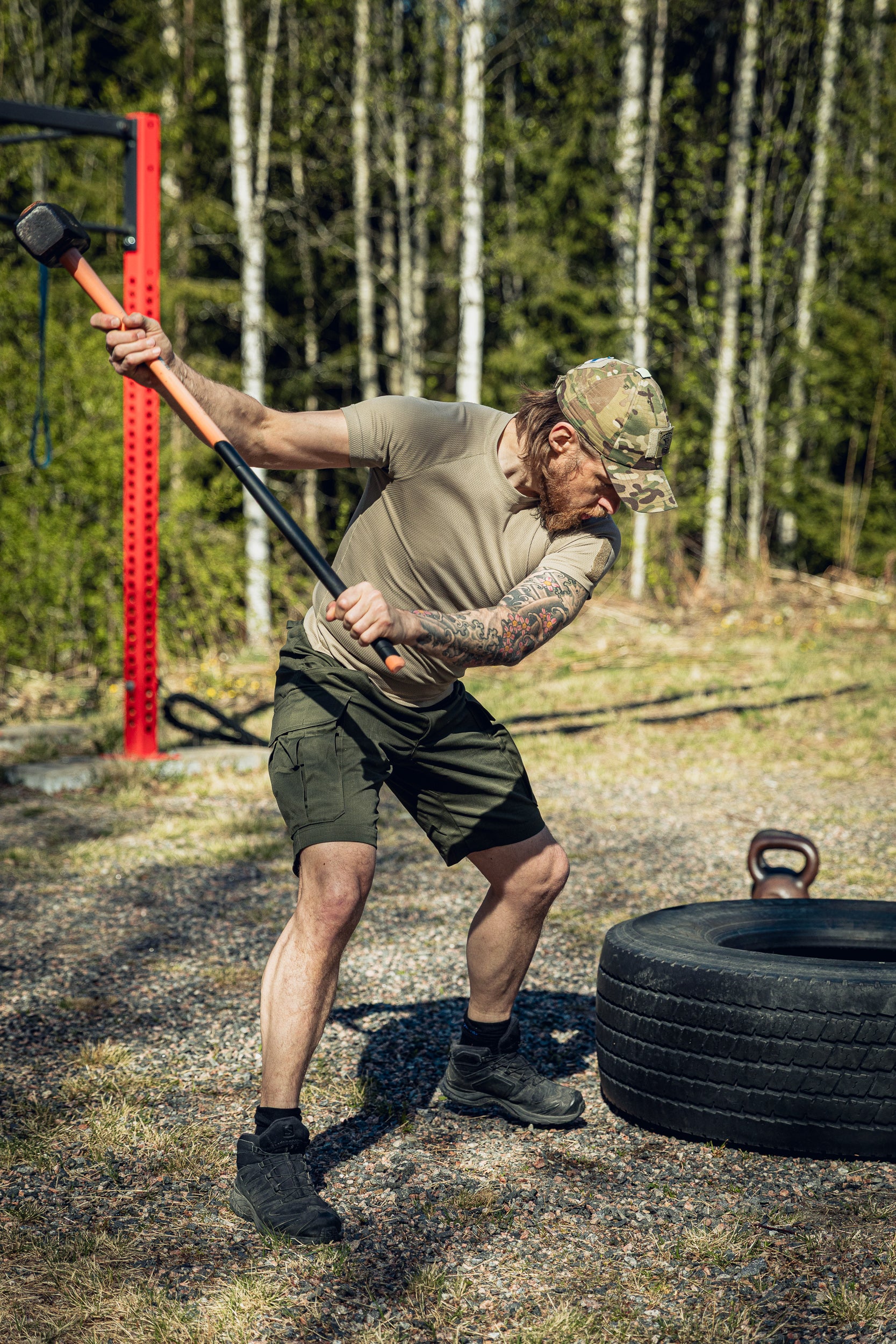 A man in a khaki t-shirt, multicam cap and green shorts hitting a tractor tire with a sledgehammer at an outdoor gym.
