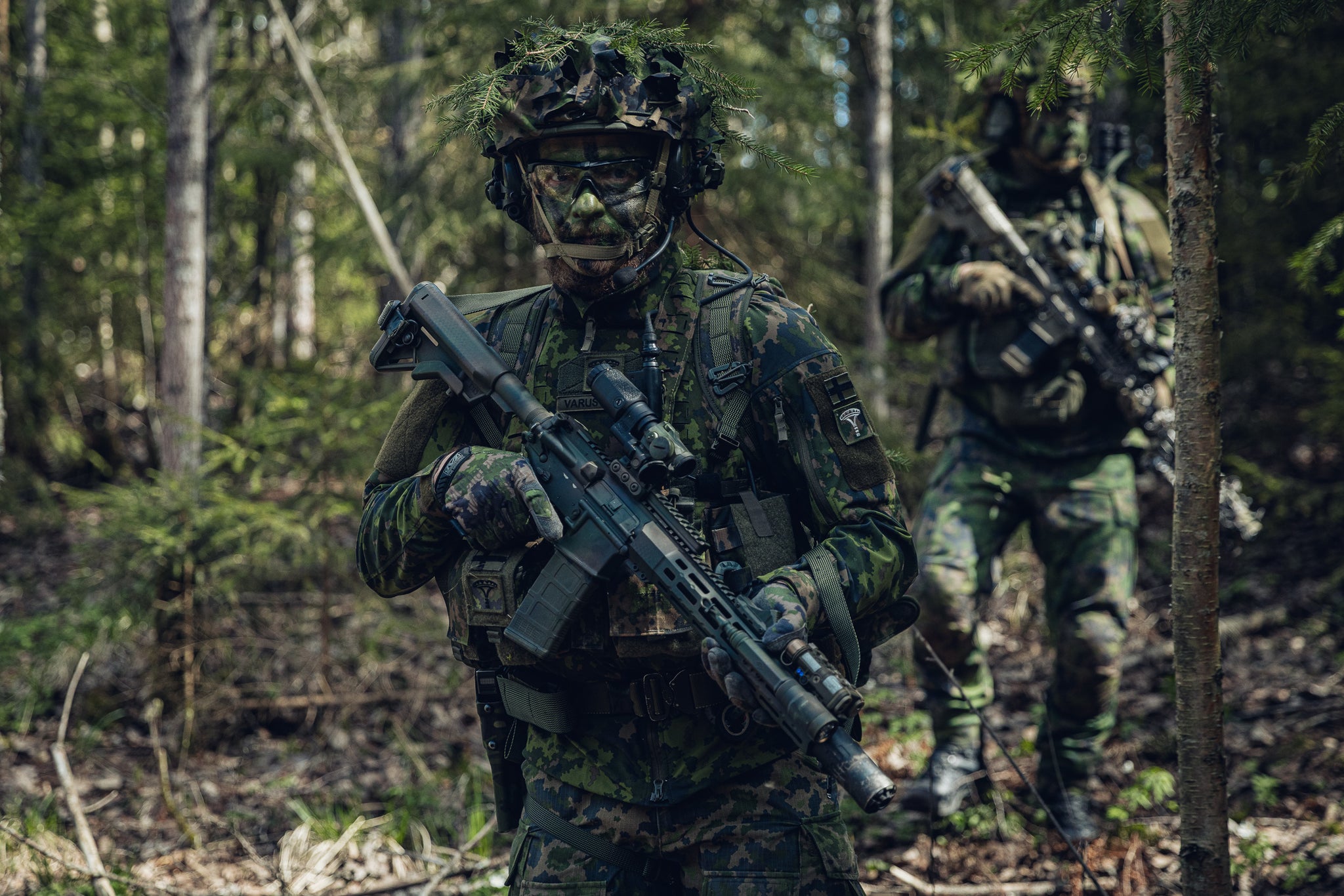 Two soldiers in Finnish woodland camouflage uniforms and helmets with foliage netting stand in a forest. The foreground soldier holds a black tactical rifle with a scope, while another soldier is visible in the blurred background.