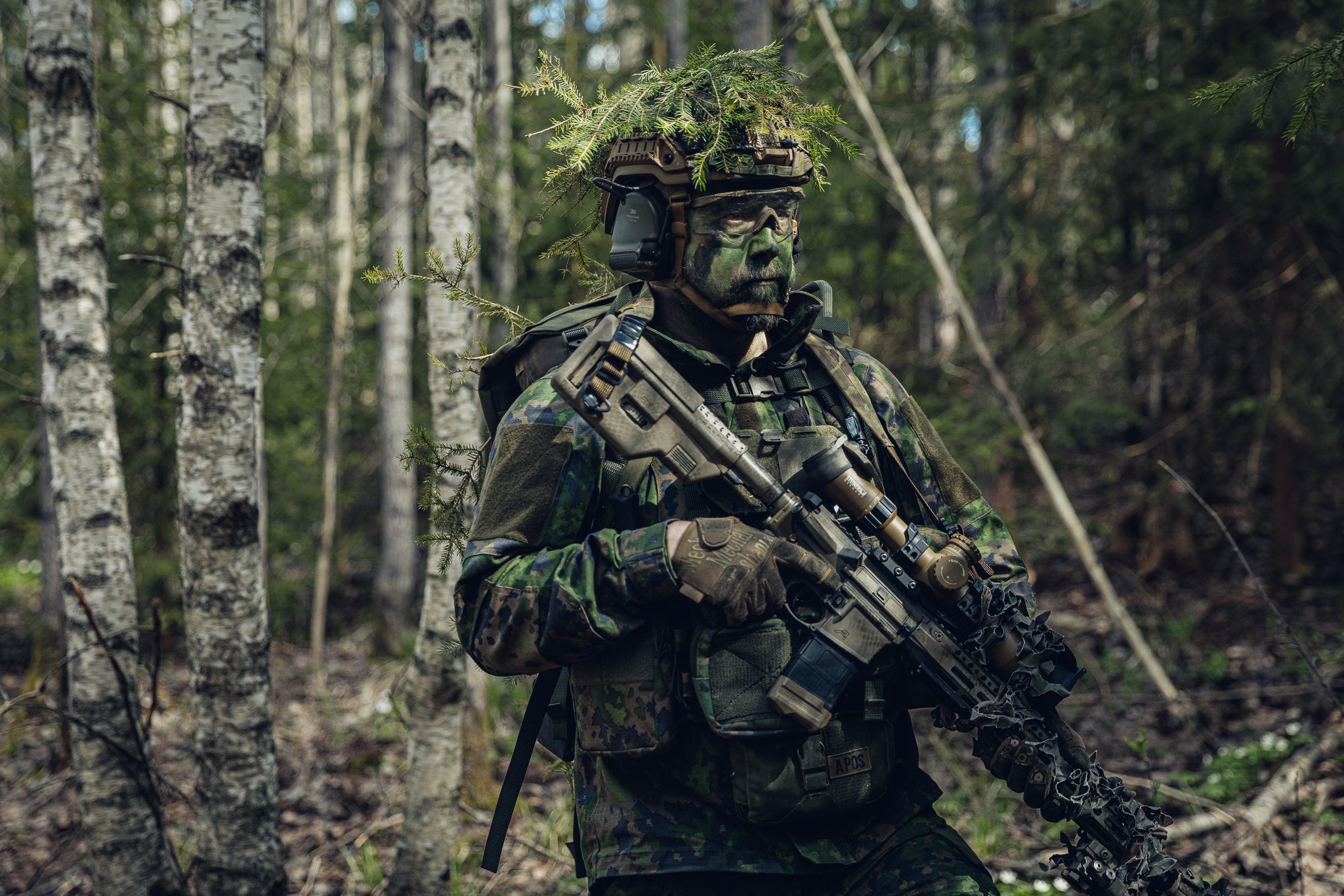 A person in full Finnish M05 camouflage military-style gear, including face paint, a helmet covered in foliage, a plate carrier and a rifle, standing in a forest among birch trees.