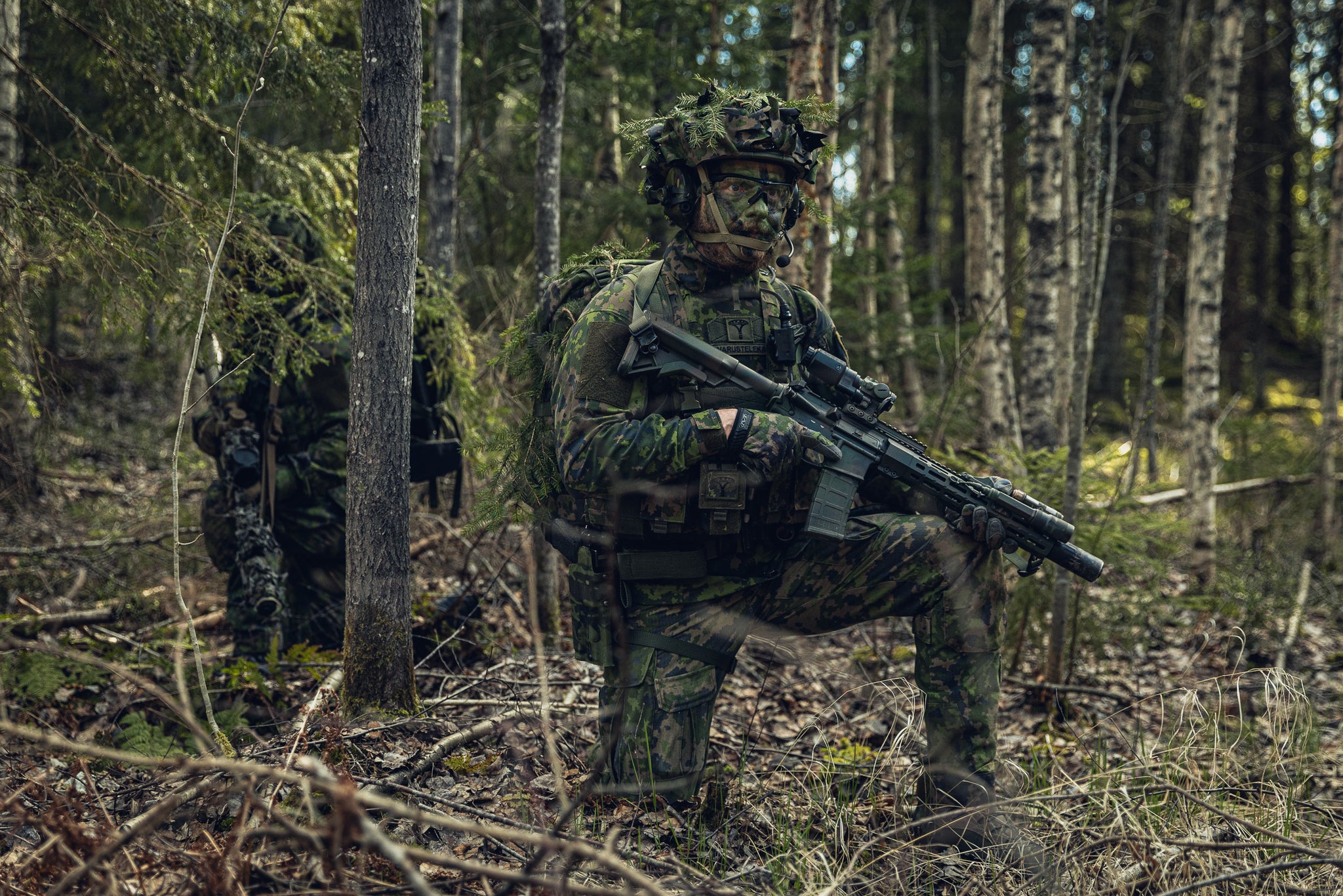 Two soldiers in full Finnish M05 camouflage military gear, crouching in a dense forest while carrying rifles.