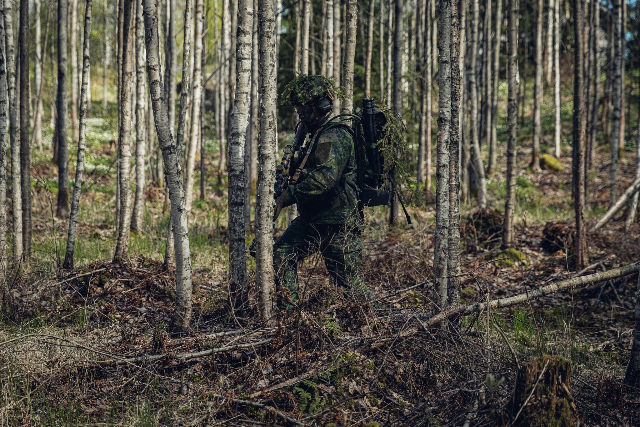A man in Finnish M05 camo gear holding a rifle moving through a birch forest.