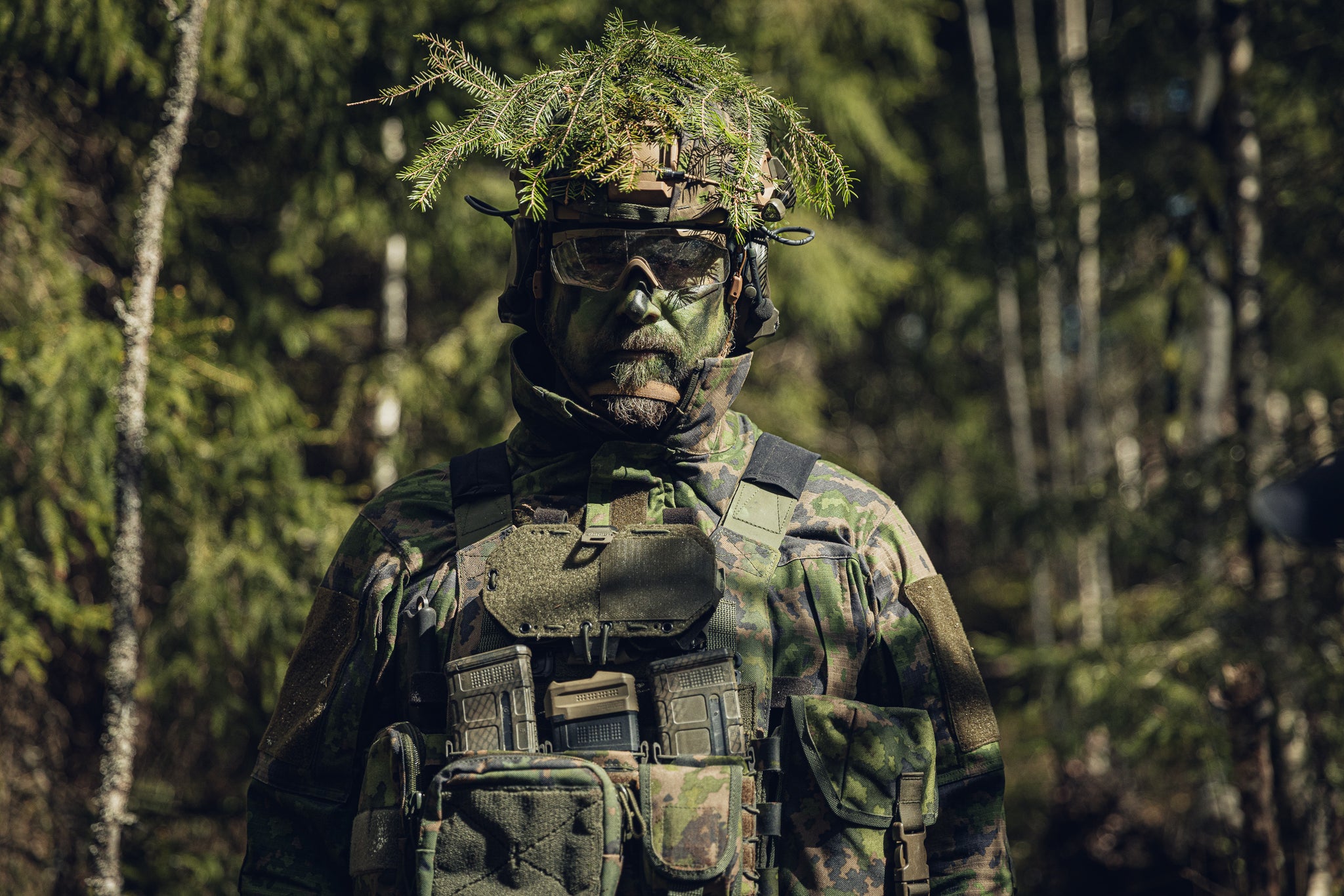A person in full military-style camouflage gear stands in a forest, wearing a helmet with leafy camouflage netting, protective goggles, and a plate carrier with pouches.