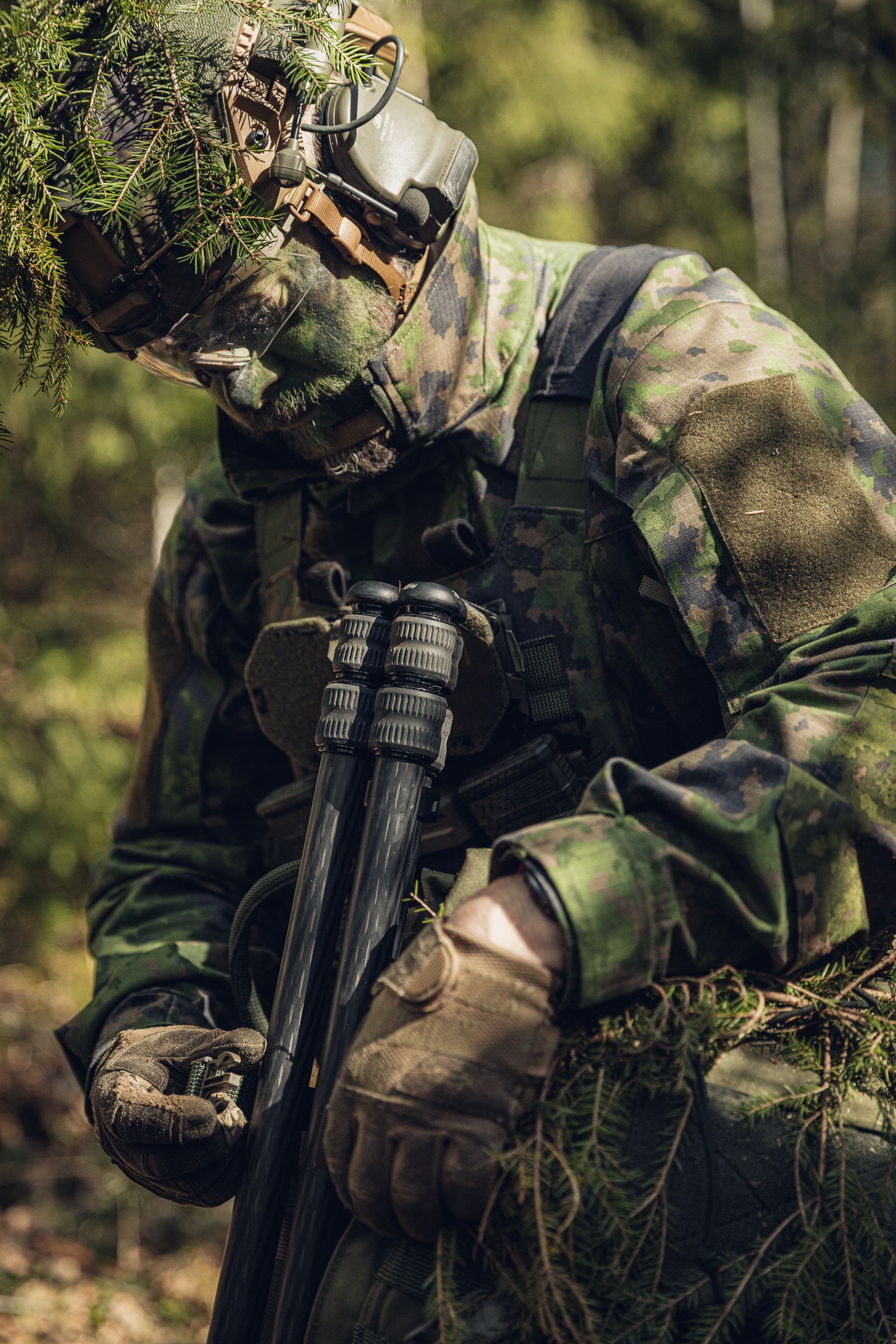 A person in full camouflage military-style gear, including a helmet with leafy ghillie netting, face covering, gloves, and a tactical vest, attaching a tripod to his backpack in a forest setting.