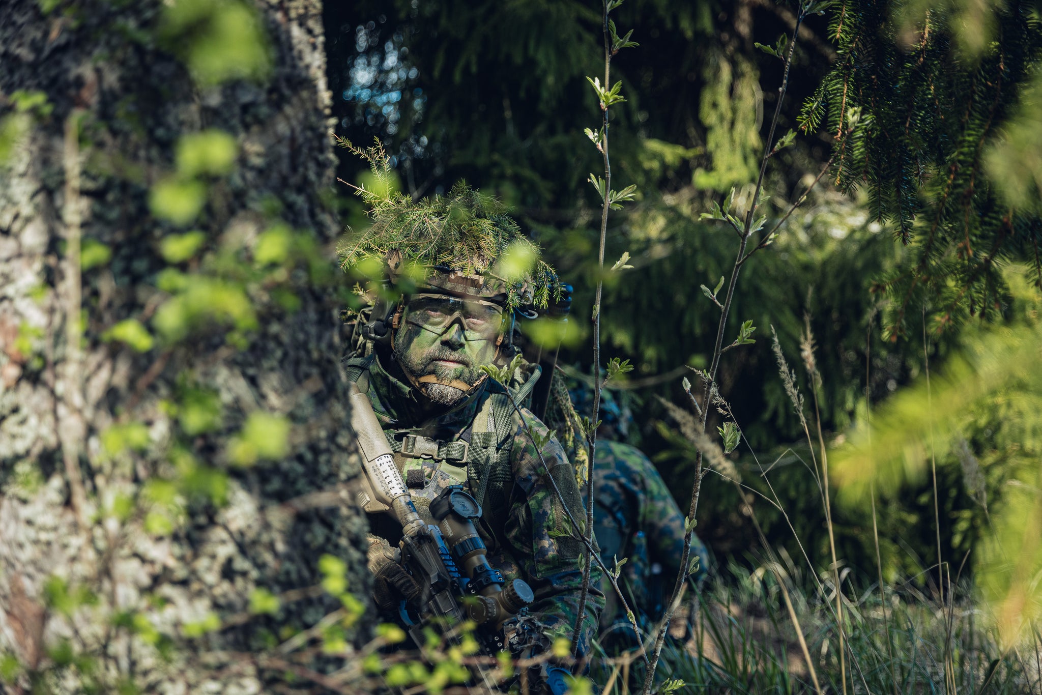 A person in full camouflage clothing and a leafy camouflage head covering crouches in a dense forest, holding a rifle. The scene is heavily obscured by foreground foliage and tree bark, set in a natural outdoor environment.