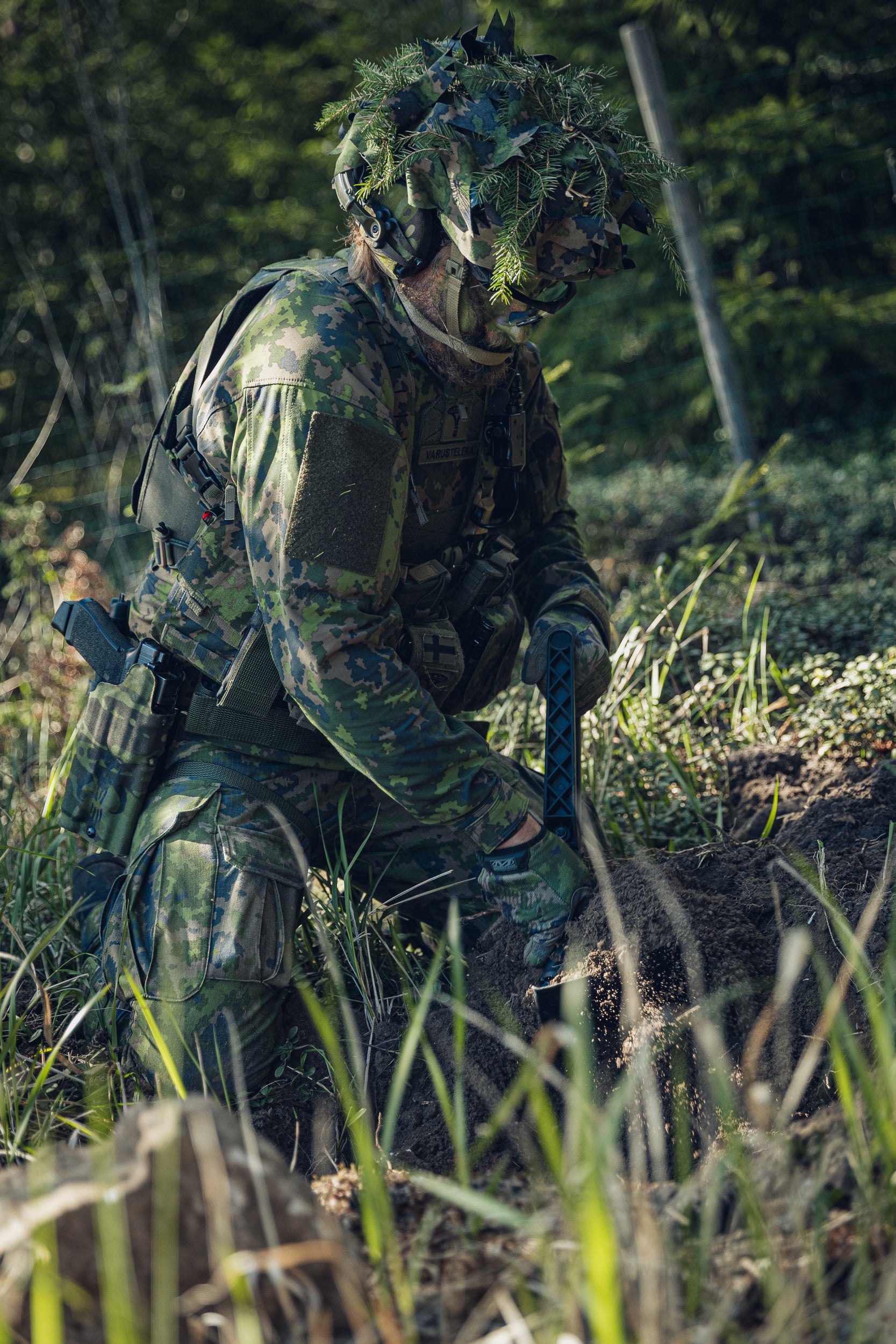 A person in full Finnish camouflage military-style gear, including a leafy helmet and face paint, kneeling on the ground, digging a hole in a forest.